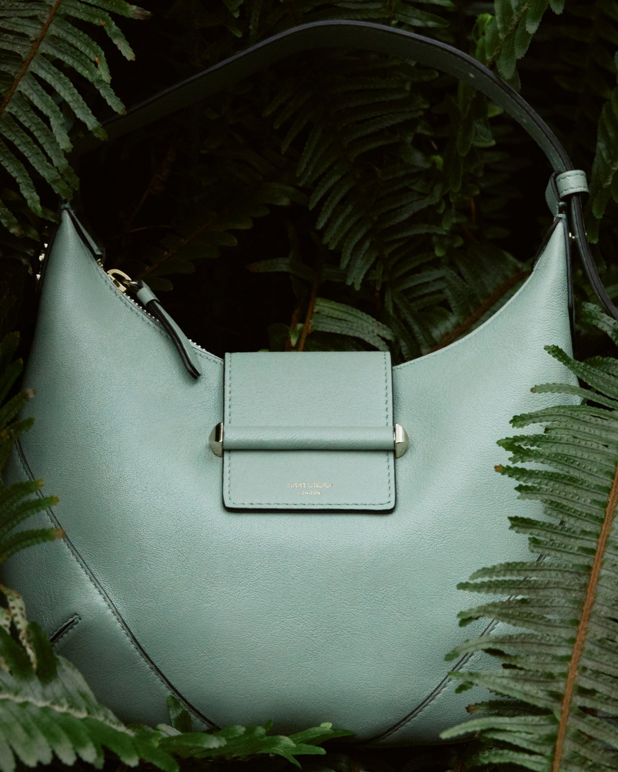 Sage green leather shoulder bag with flap detail, styled among fern leaves.