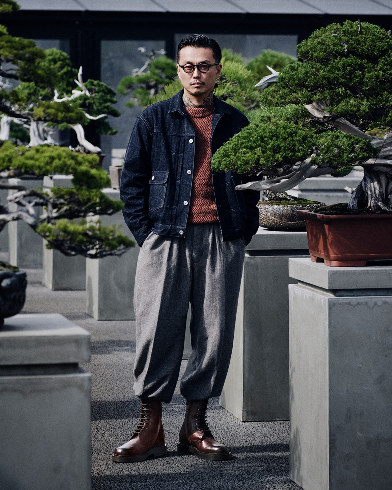A man wearing a jacket and trousers stands among bonsai trees arranged on concrete plinths, wearing dark leather boots.