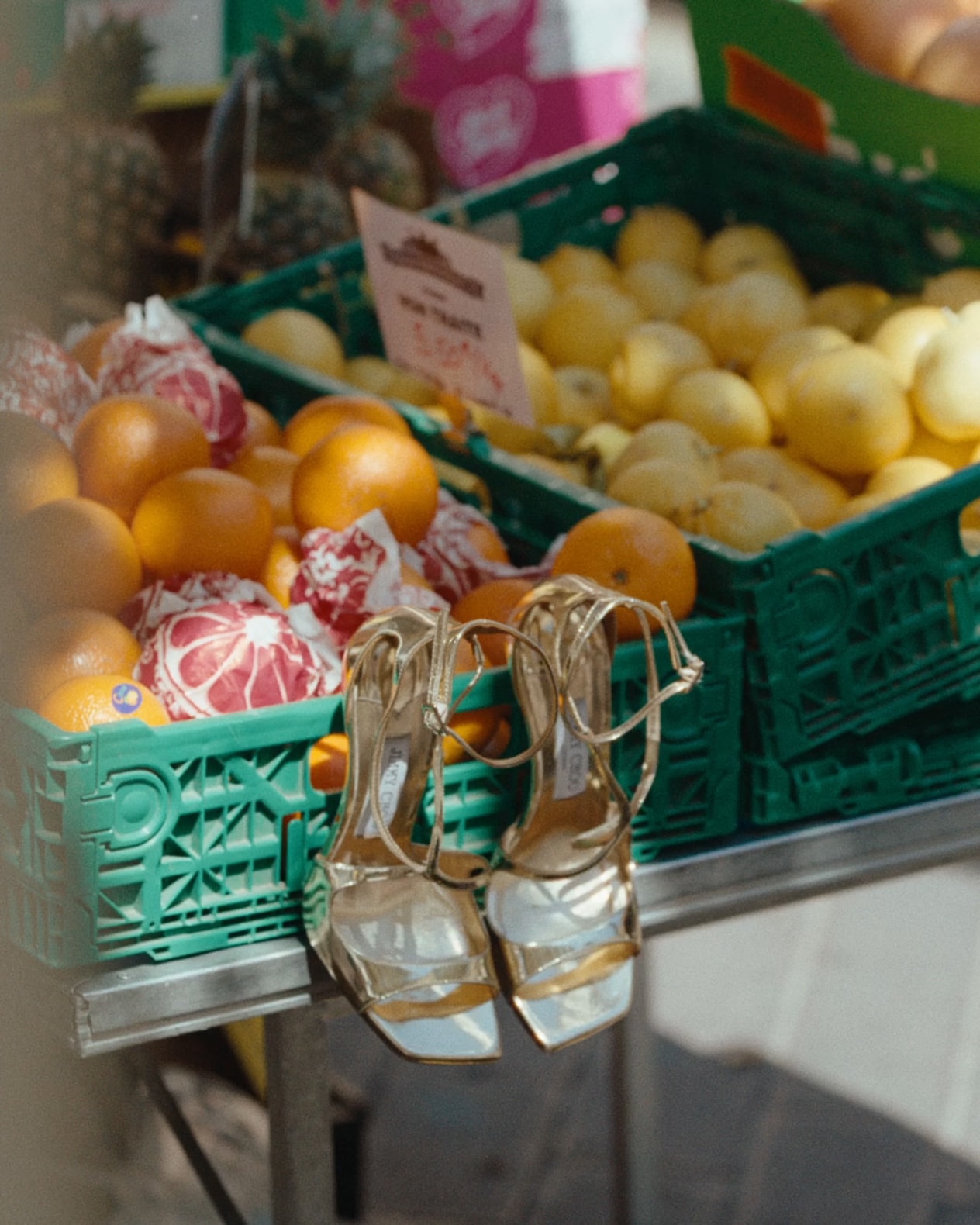 Pair of gold sandals displayed in front of crates of citrus fruit at an outdoor market.