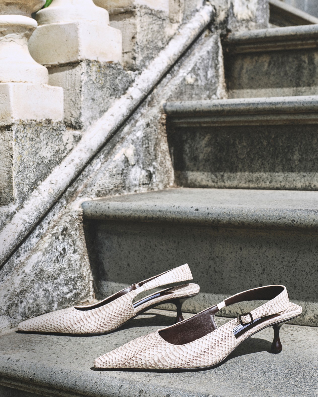 Close-up of a womans legs crossed while wearing strappy beige heeled sandals with pearl embellishments, resting on cream-coloured bedding.