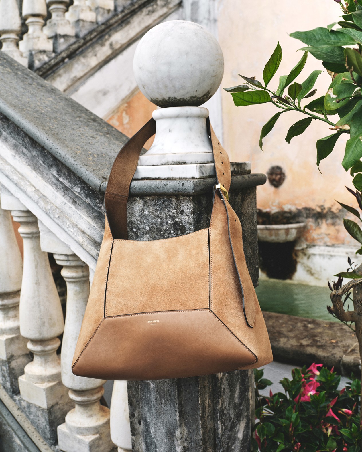 Beige drawstring shoulder bag with dark brown floral embroidery, gold hardware and circular top handle, displayed on a gold door handle against a paneled wall.