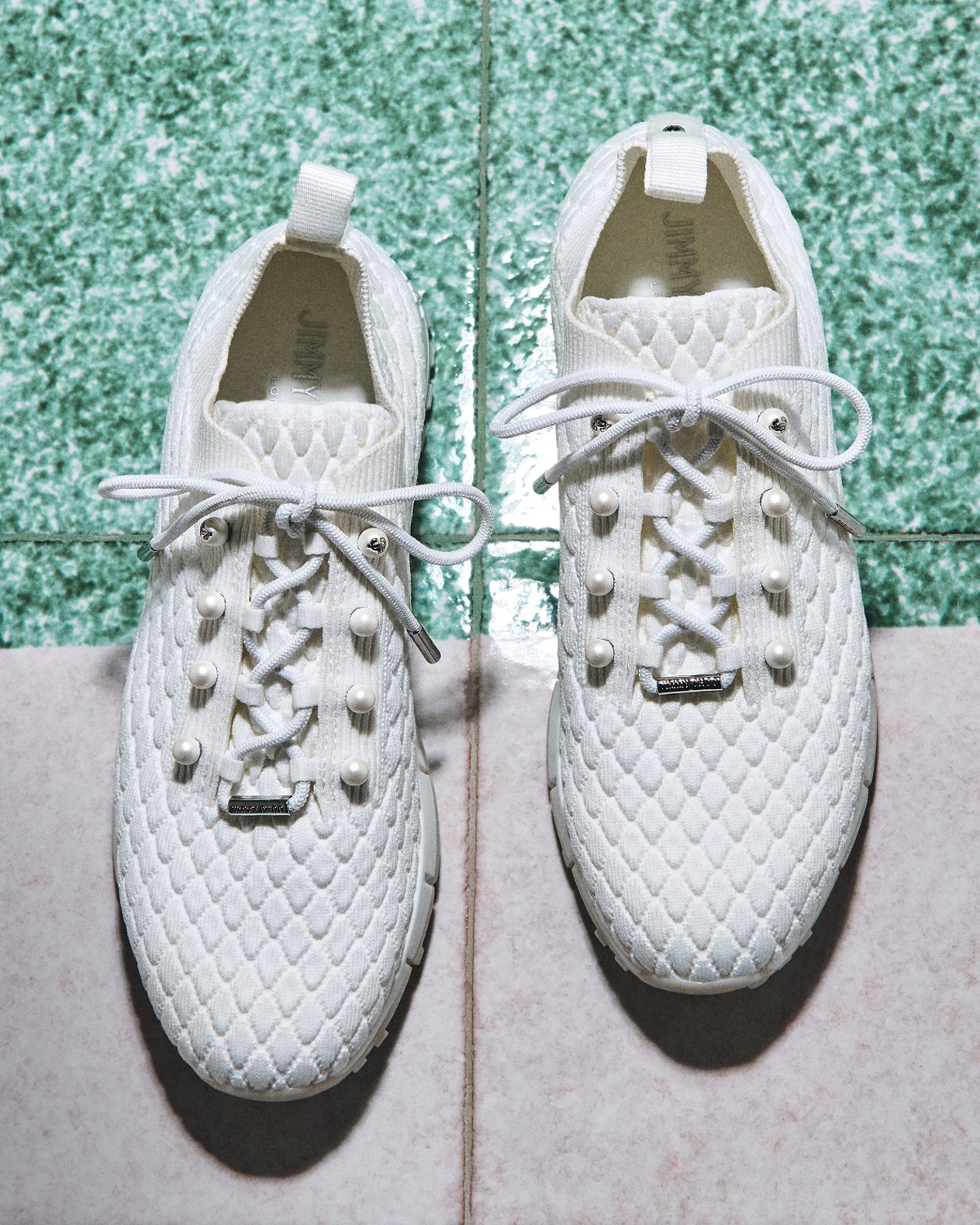 Close-up of a womans legs crossed while wearing strappy beige heeled sandals with pearl embellishments, resting on cream-coloured bedding.