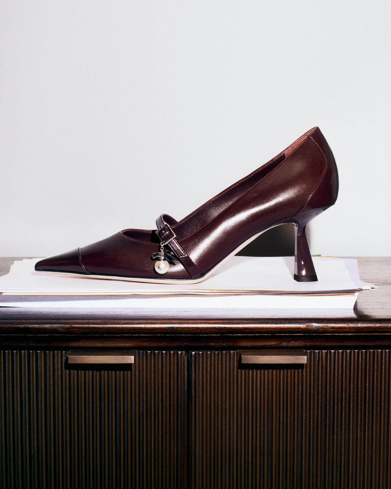 Brown leather pointed-toe pump with a sleek micro heel and silver buckle and pearl charm, displayed on stacked papers against a minimalist background.