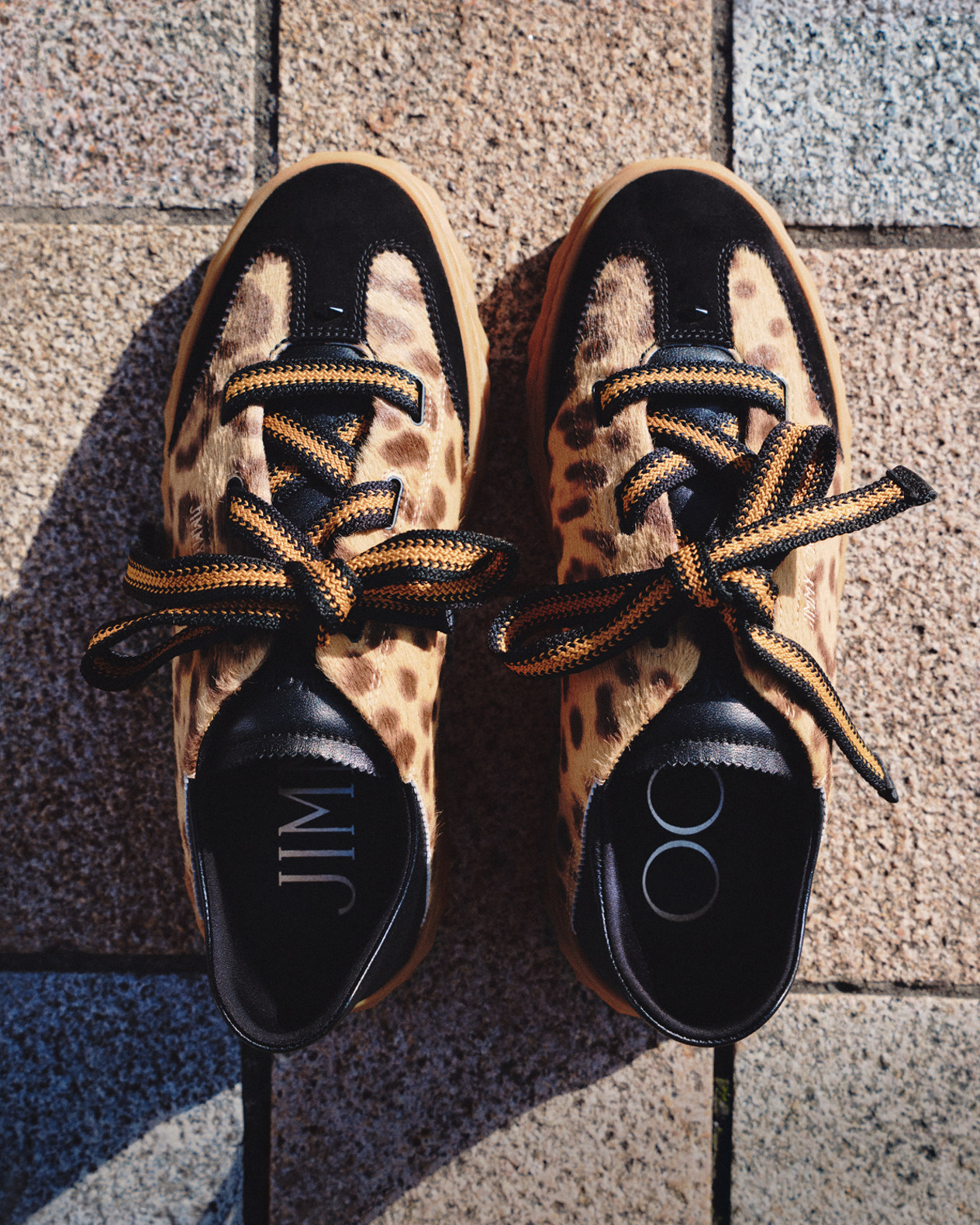 Pair of cheetah-print trainers with leather trim, featuring oversized laces, photographed on a stone surface.