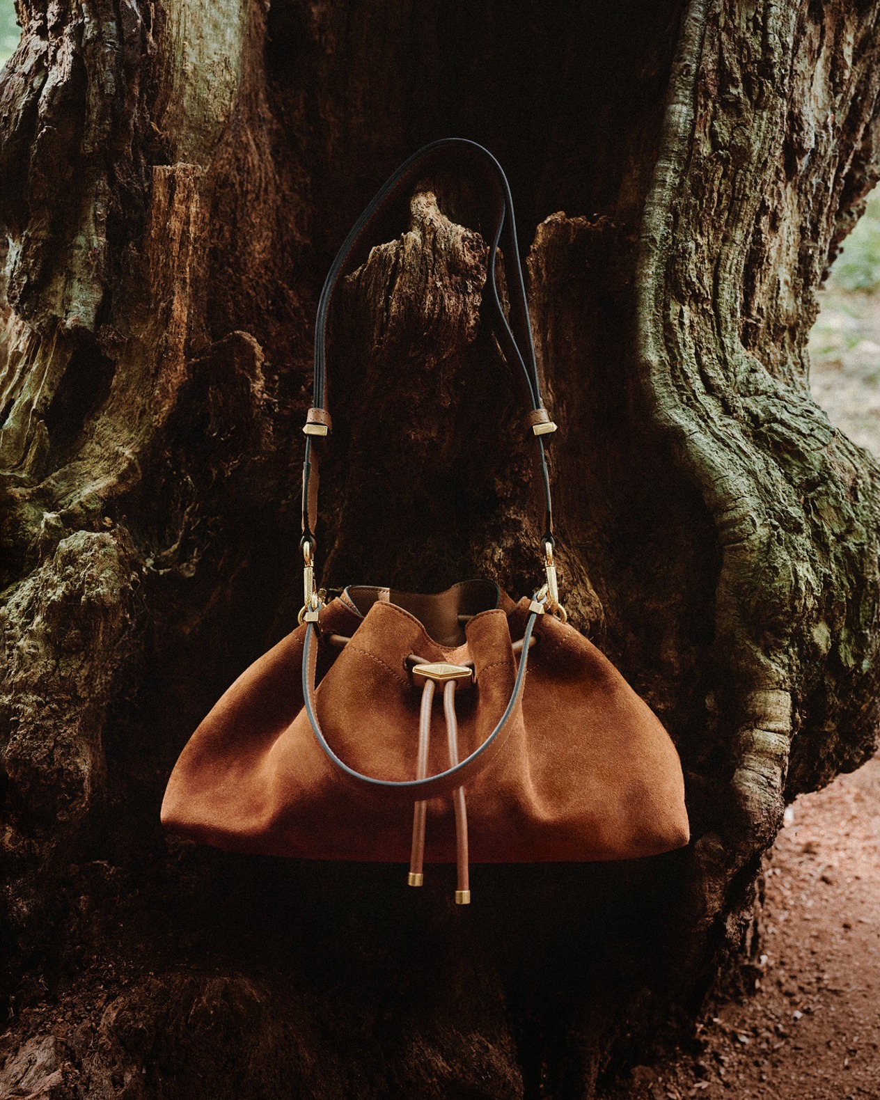 Cream knitted drawstring shoulder bag with leather strap, styled against textured tree bark in a natural forest setting. 