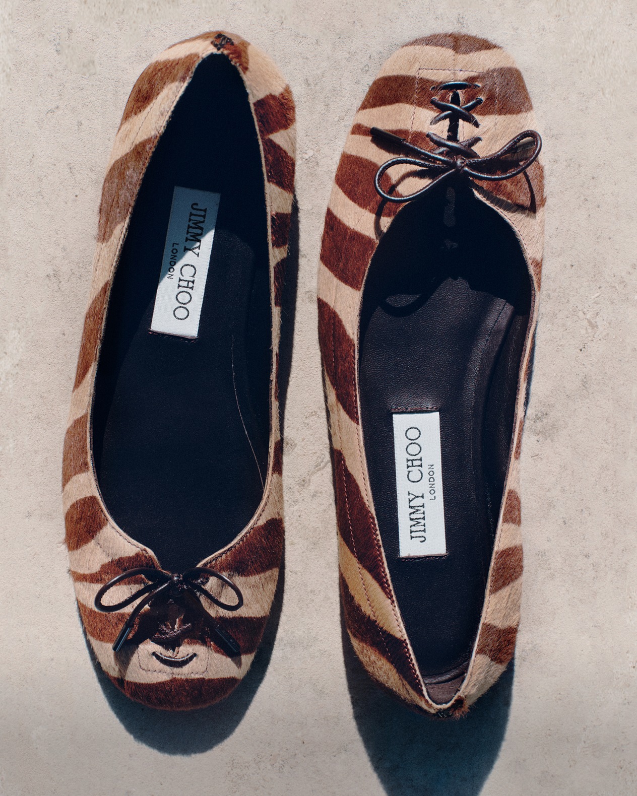 Brown leather pointed-toe pump with a sleek micro heel and silver buckle and pearl charm, displayed on stacked papers against a minimalist background.