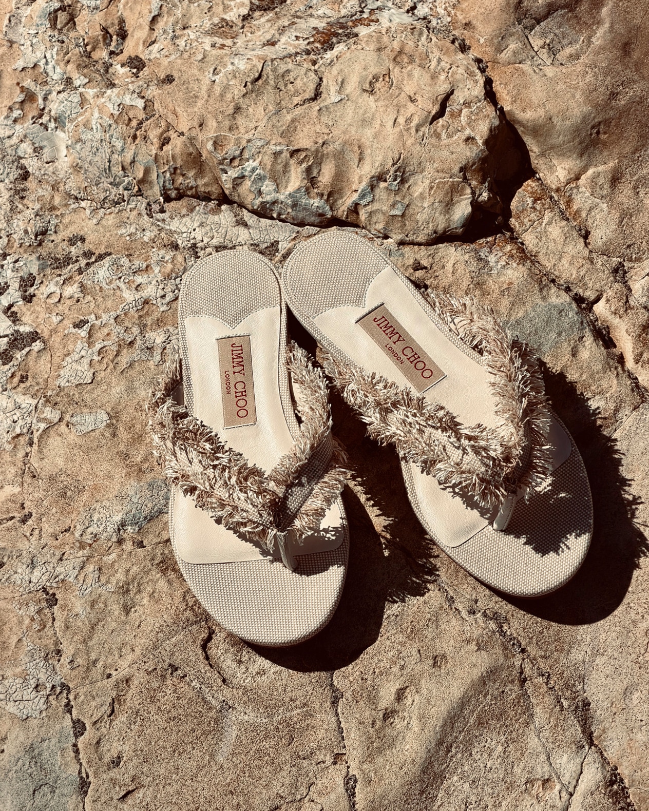 Beige flat sandals with textured fringe straps placed on rocky ground.