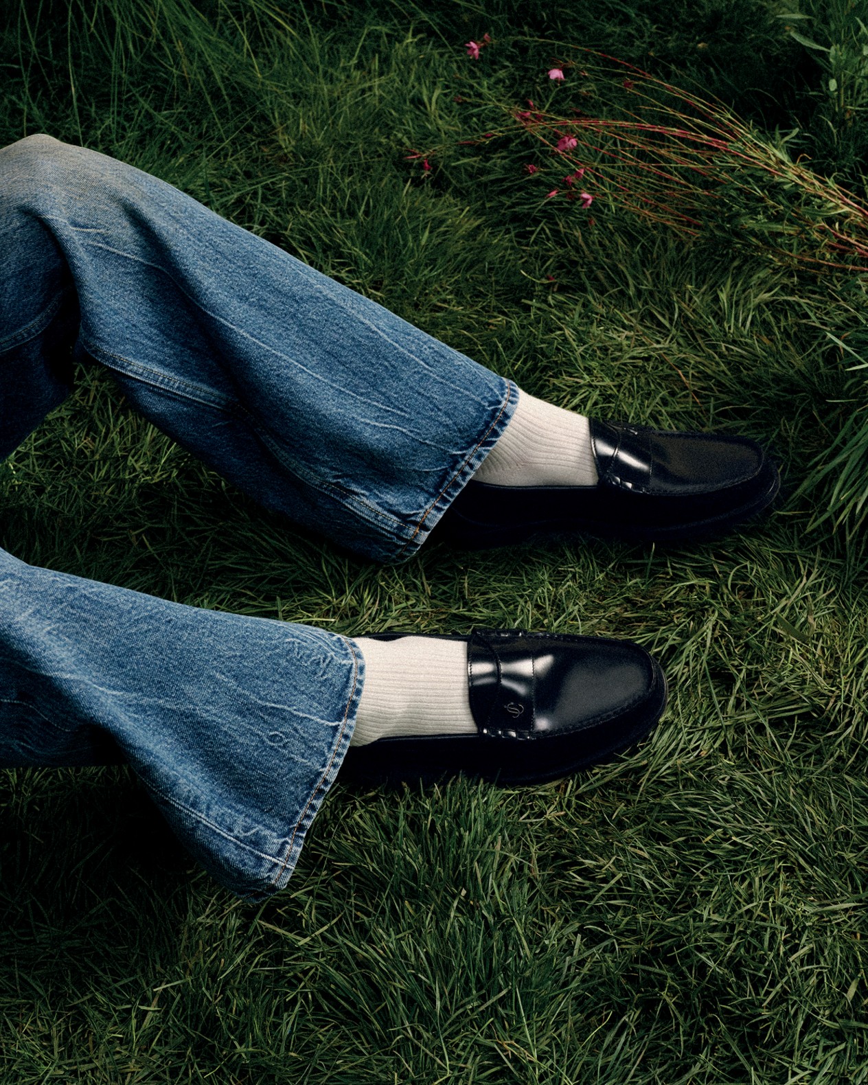 Black leather loafers worn with white socks and denim jeans on grass.