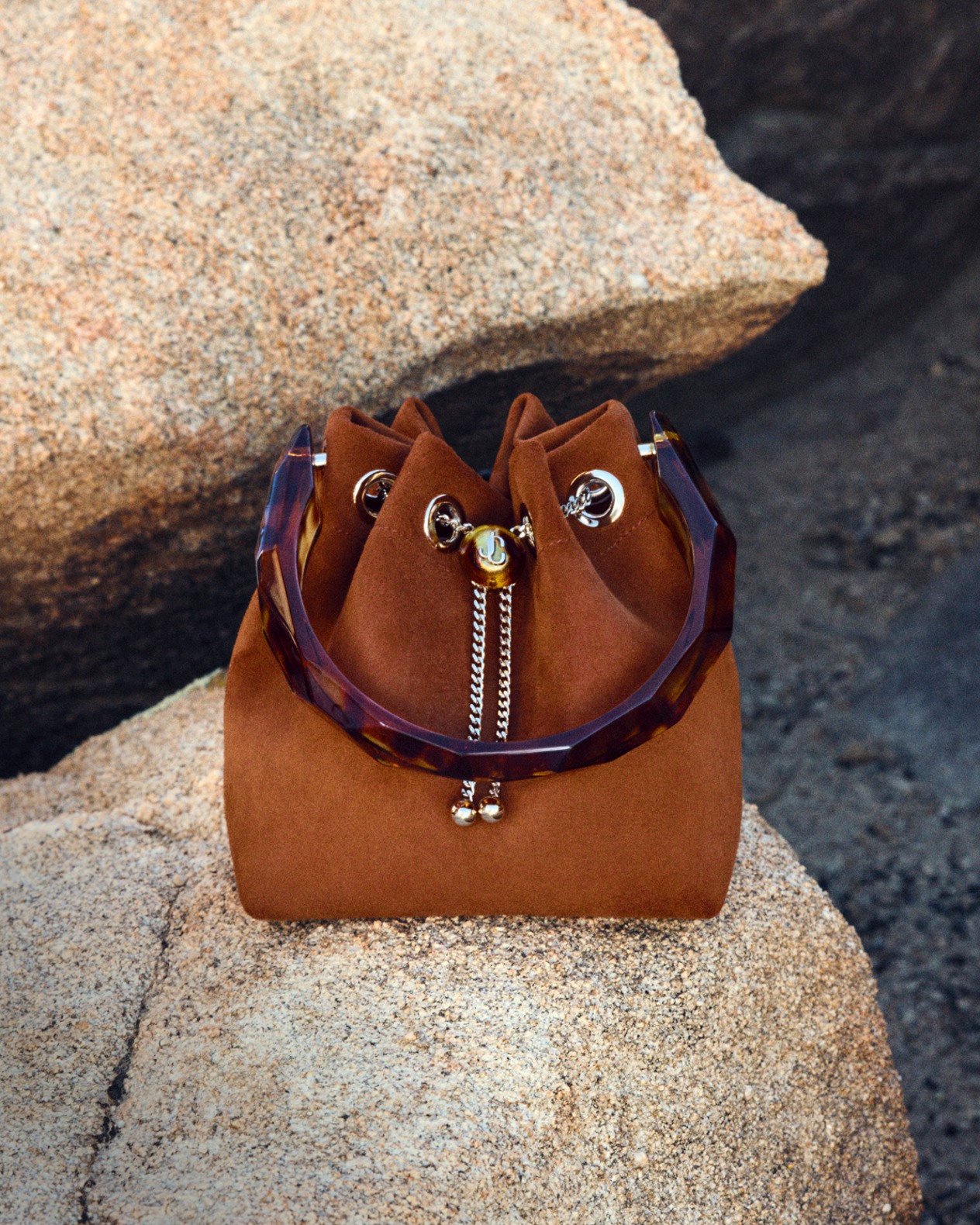 Brown suede drawstring bucket bag with tortoiseshell-effect handle detail placed on natural rocks.
