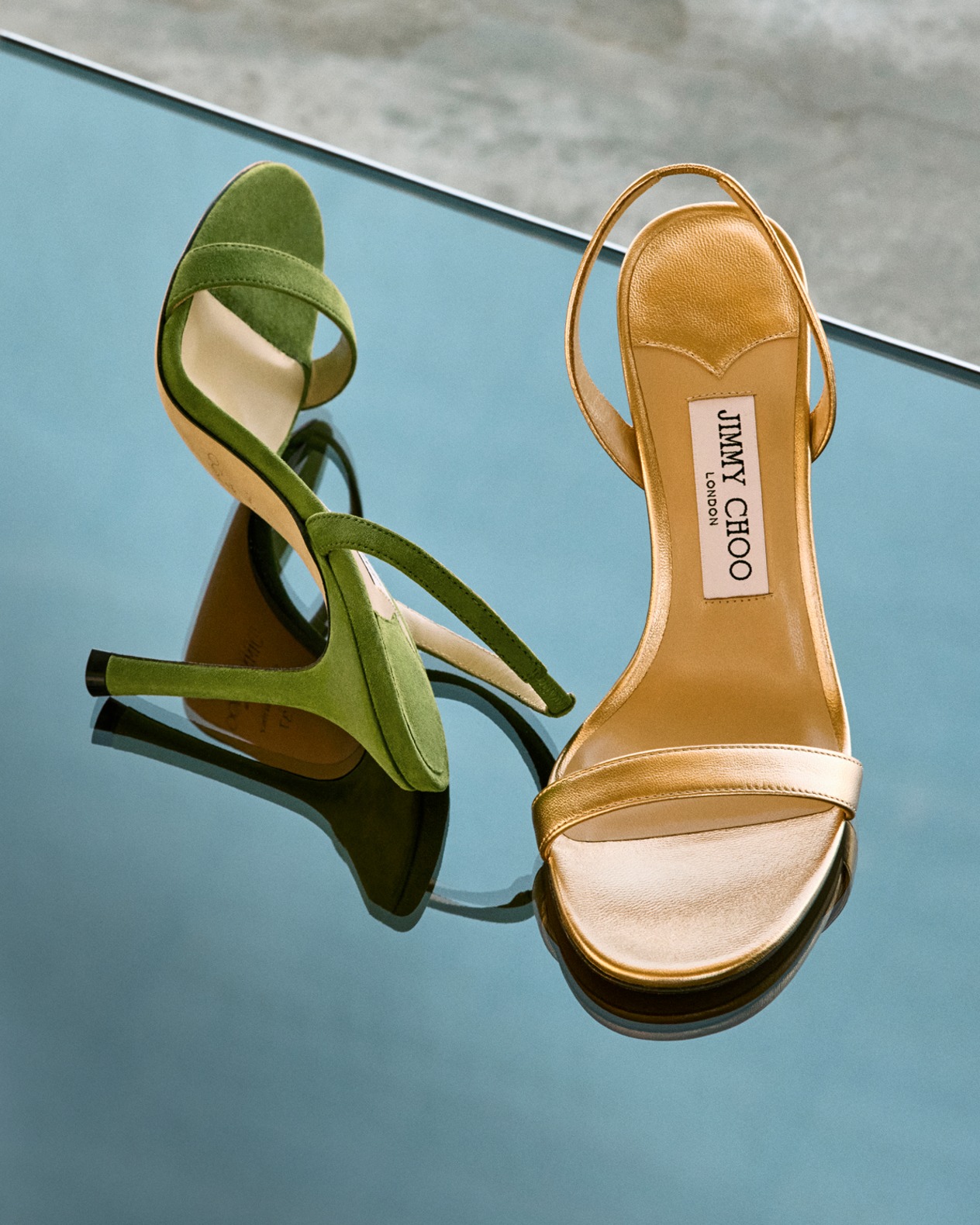 Green suede and gold leather heeled sandals displayed on a reflective blue surface.