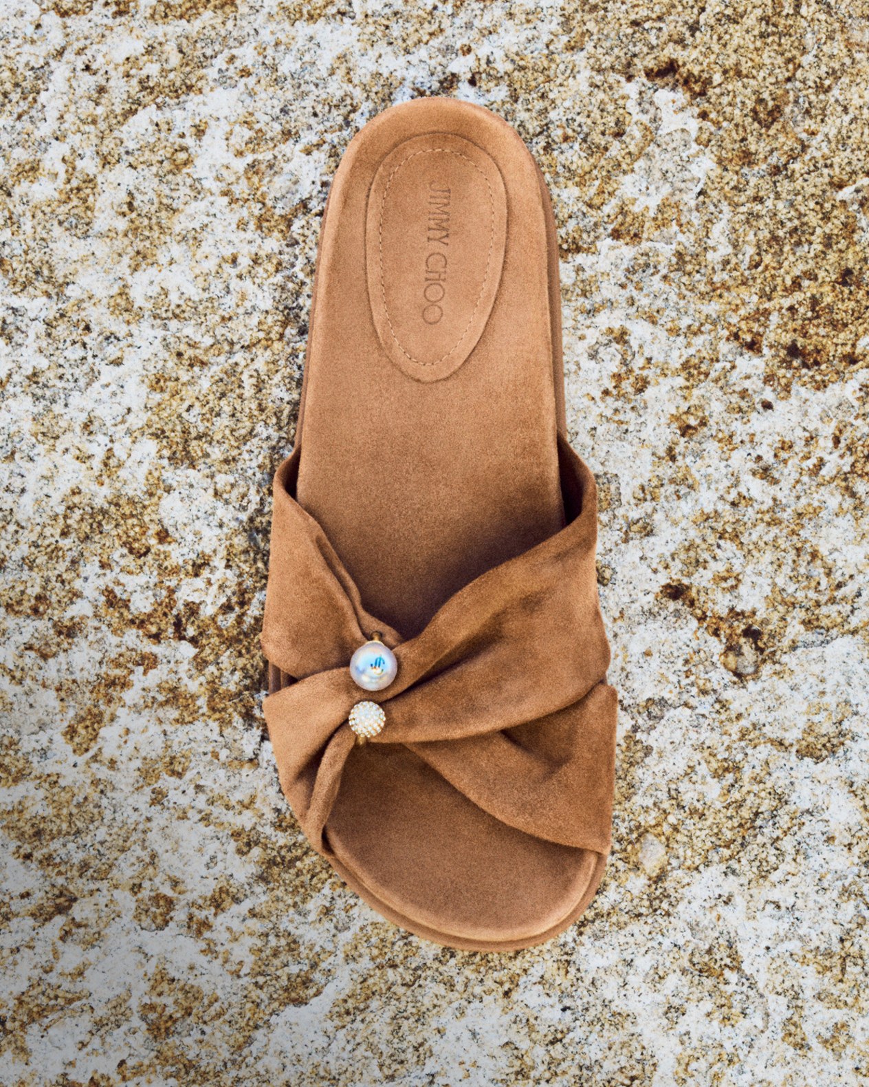 IMAGE ALT: Brown suede slide sandal with a twisted front strap and pearl and crystal embellishments displayed on a textured stone surface.