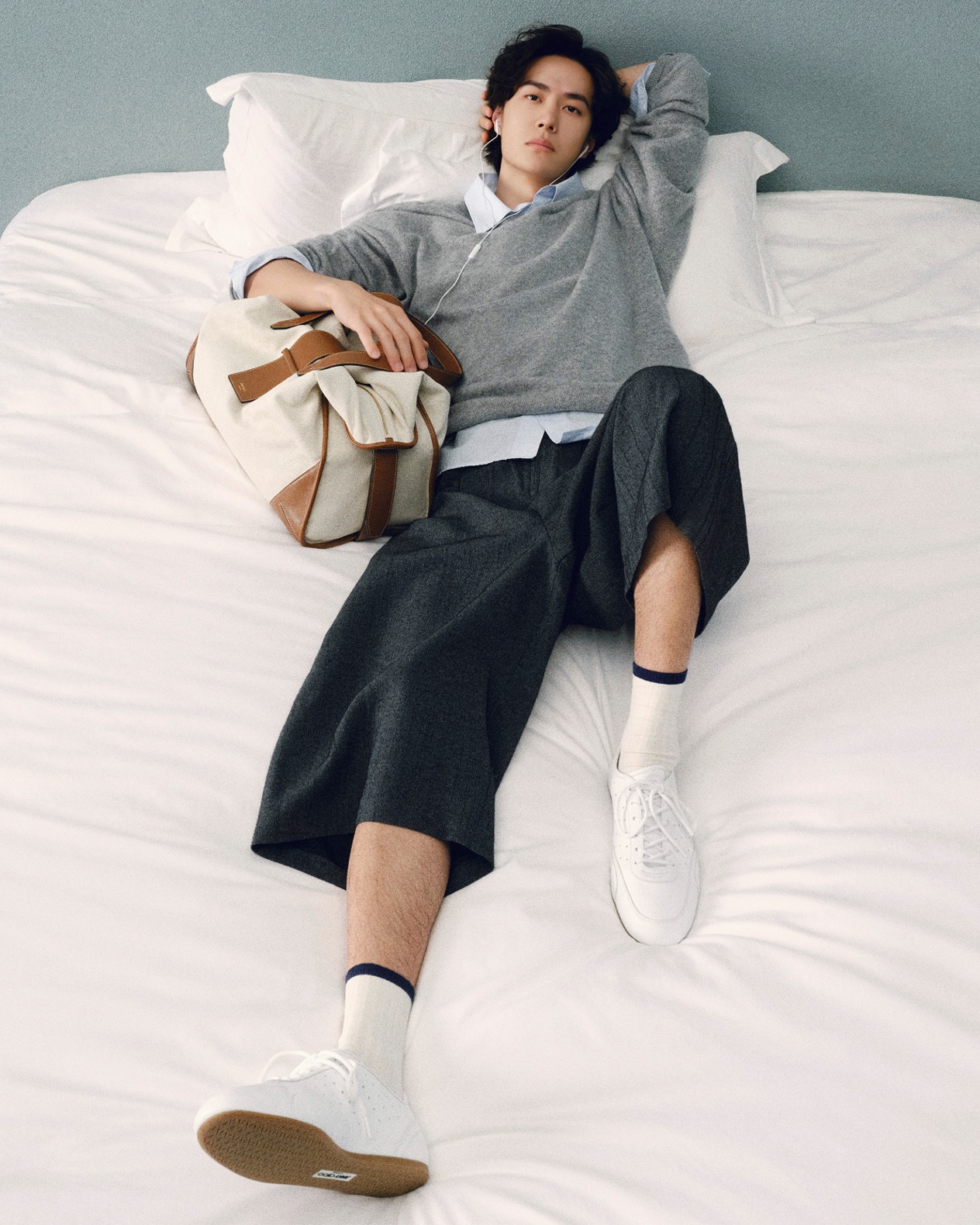 Man reclining on a white bed wearing a grey knit jumper, cropped trousers and white lace-up trainers, holding a canvas bag.			