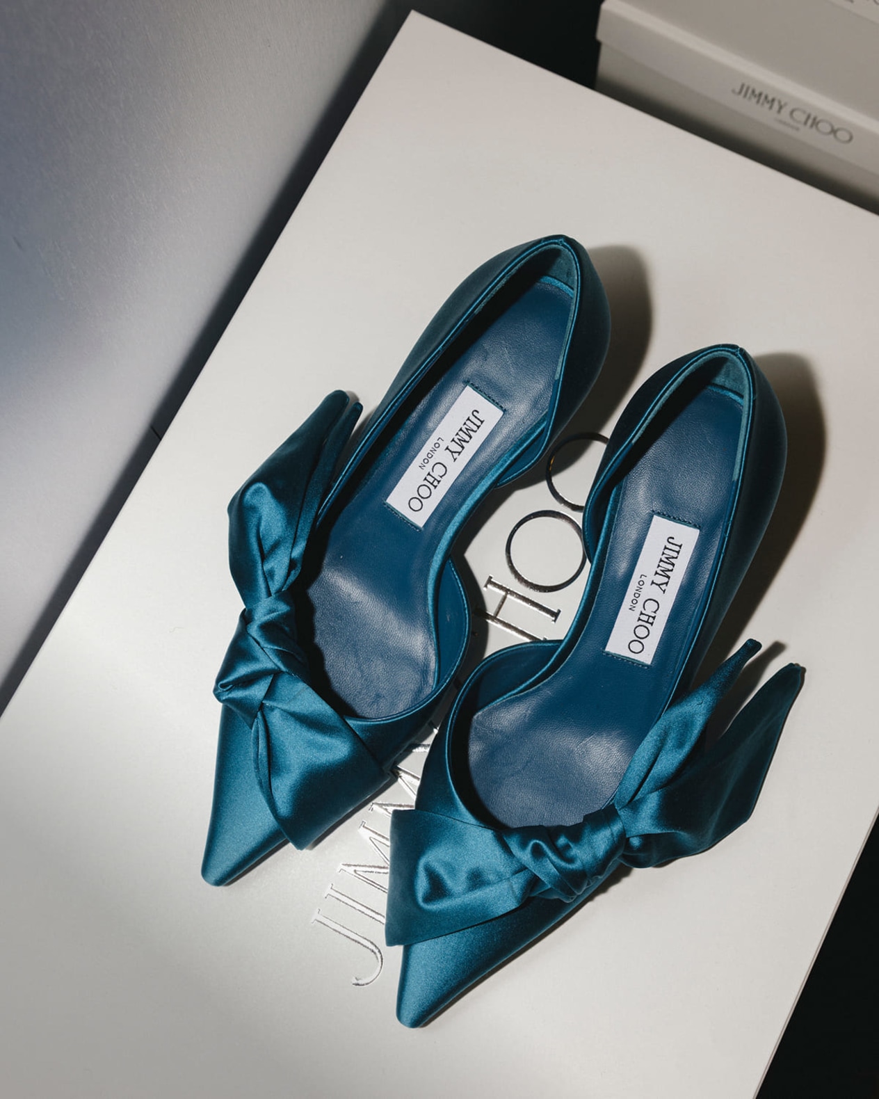 Pair of blue satin pointed-toe pumps with oversized bow detailing. Photographed on a Jimmy Choo shoe box.