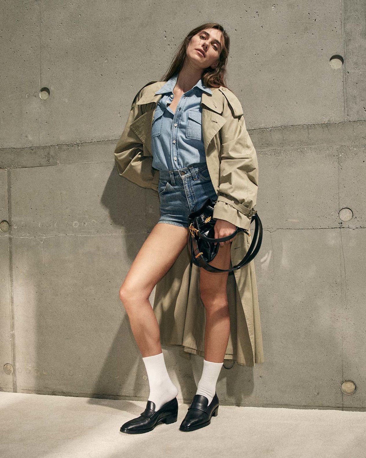 Devon Lee Carlson in a beige trench coat and denim shorts wearing black leather loafers and carrying a black patent leather drawstring bag.