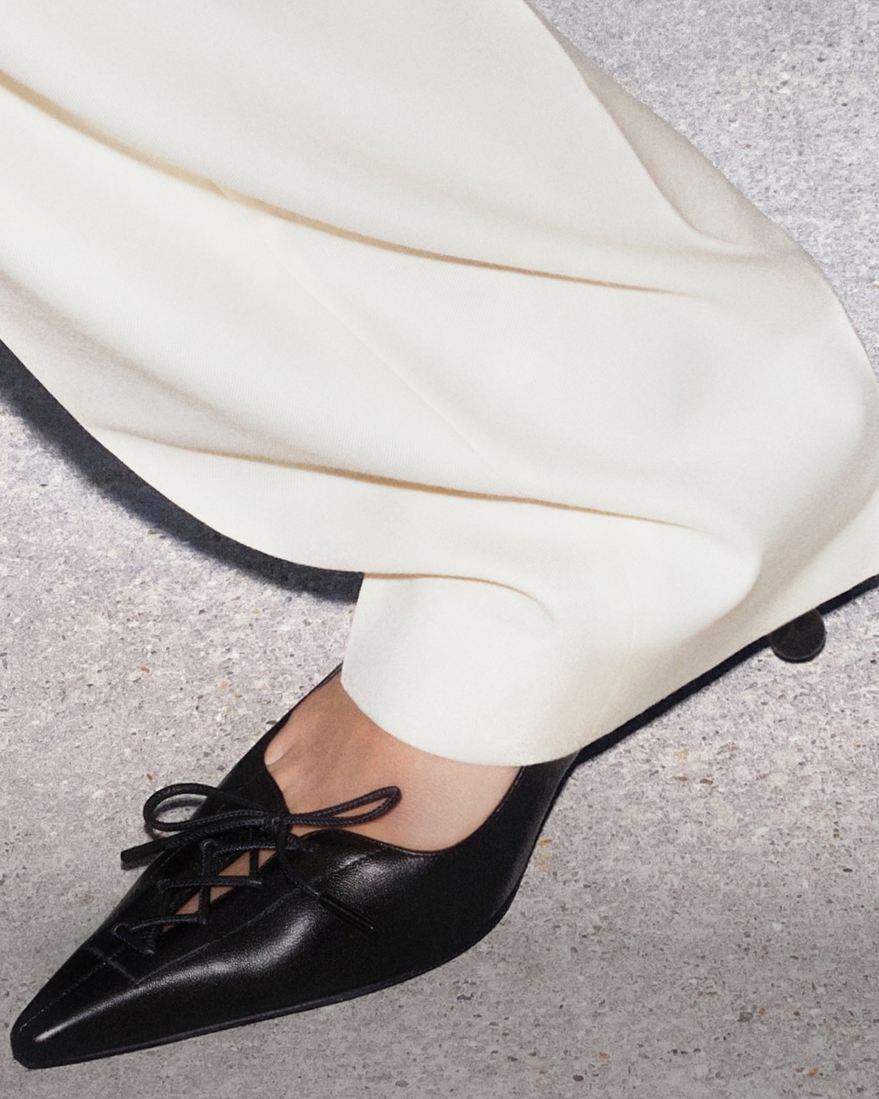 Close-up of black leather pumps with slim stiletto heels, worn with white trousers.