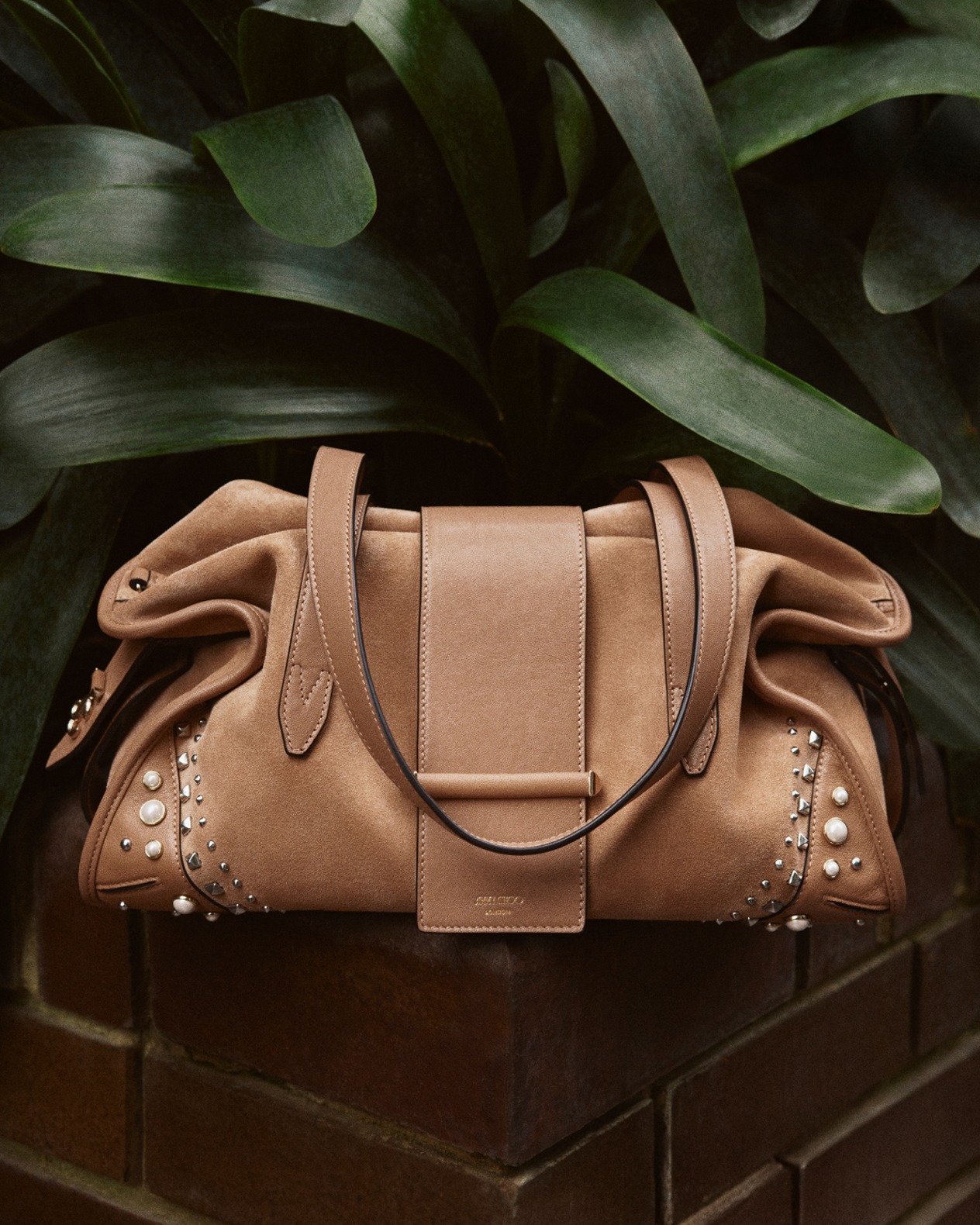 Tan leather handbag with silver stud embellishment, resting on a brick ledge against large green tropical leaves.