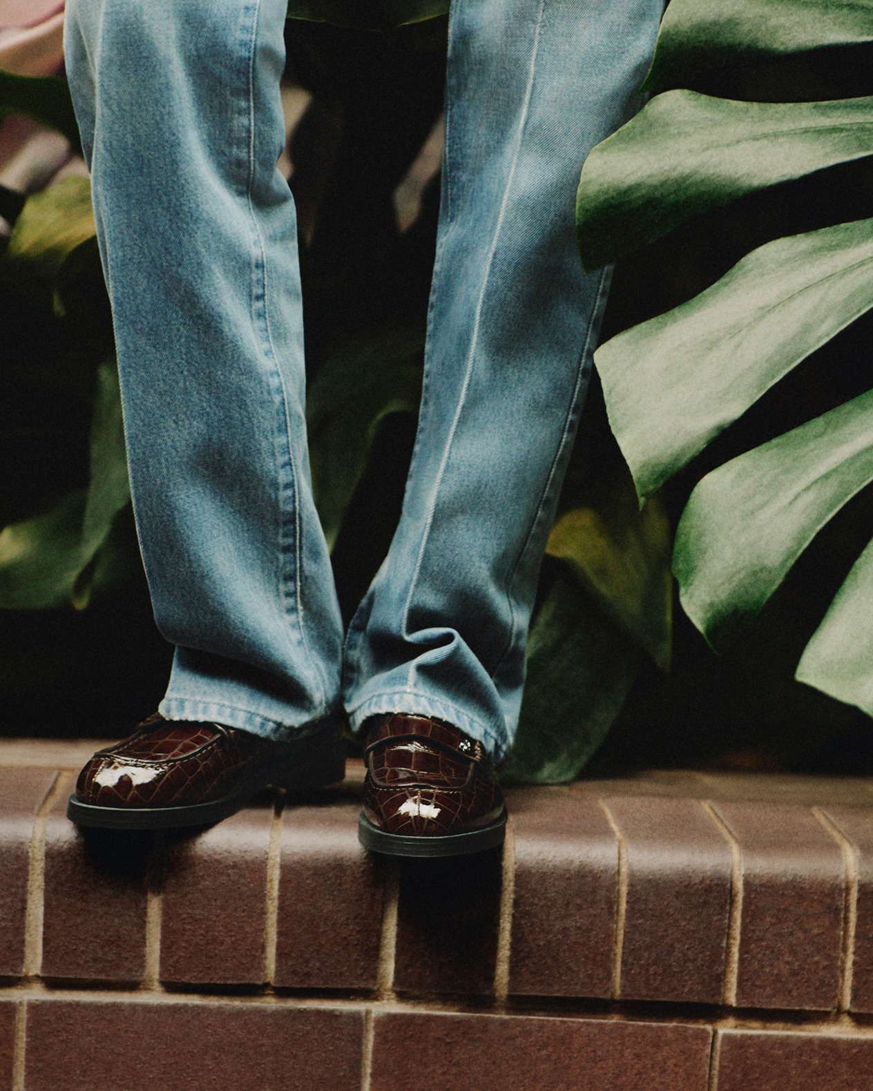 Brown leather loafers worn with straight-leg blue jeans, styled on brick steps.