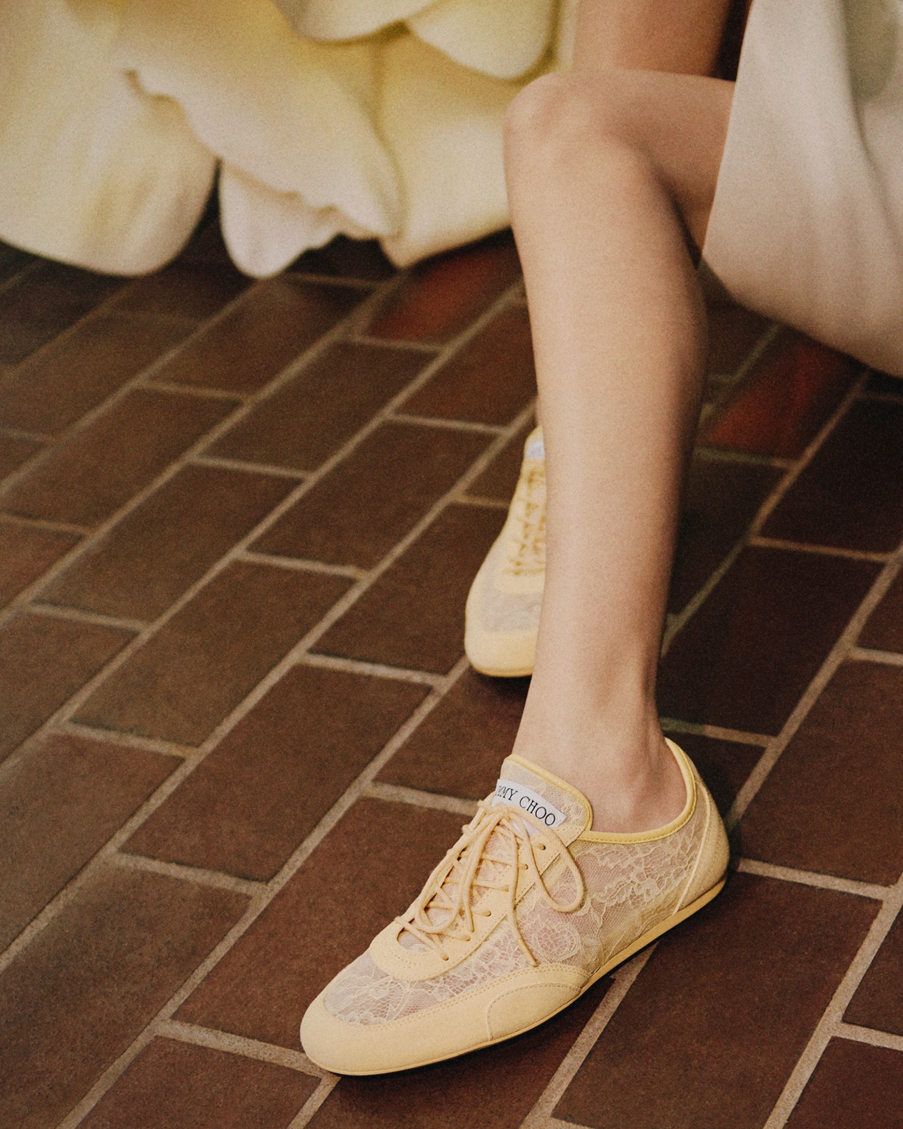 Yellow lace-up trainers with lace overlay, worn with a neutral skirt on brick flooring.