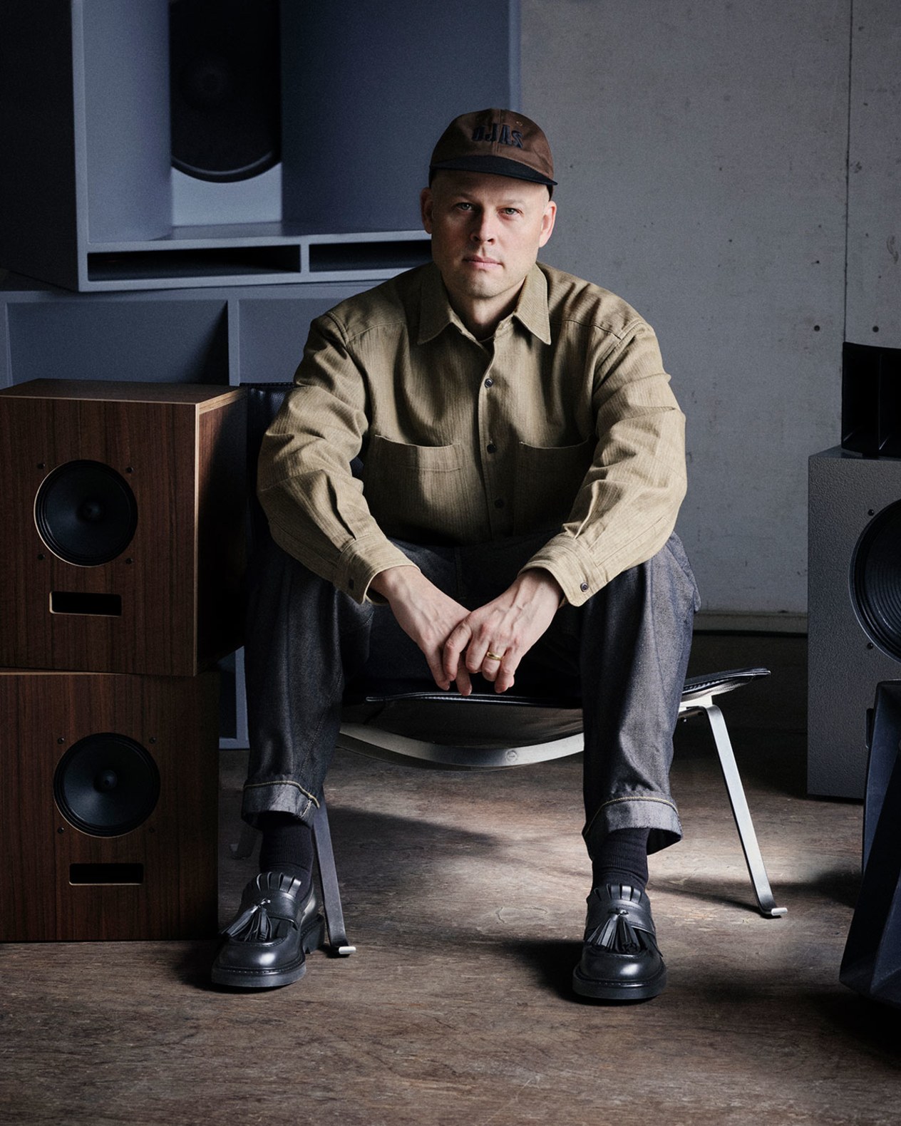 IMAGE ALT: A man wearing a brown shirt and cap sits on a low stool between large speakers, wearing black leather shoes.