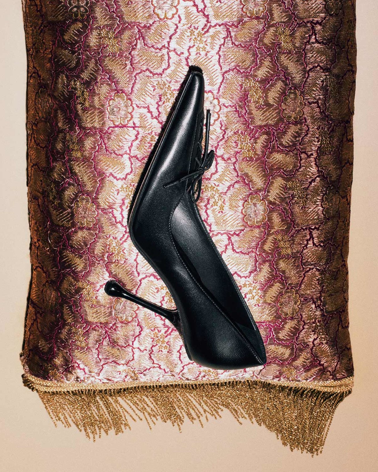 Black leather pointed-pump with a sculptural drop heel, displayed against a textured gold and burgundy fabric backdrop.
