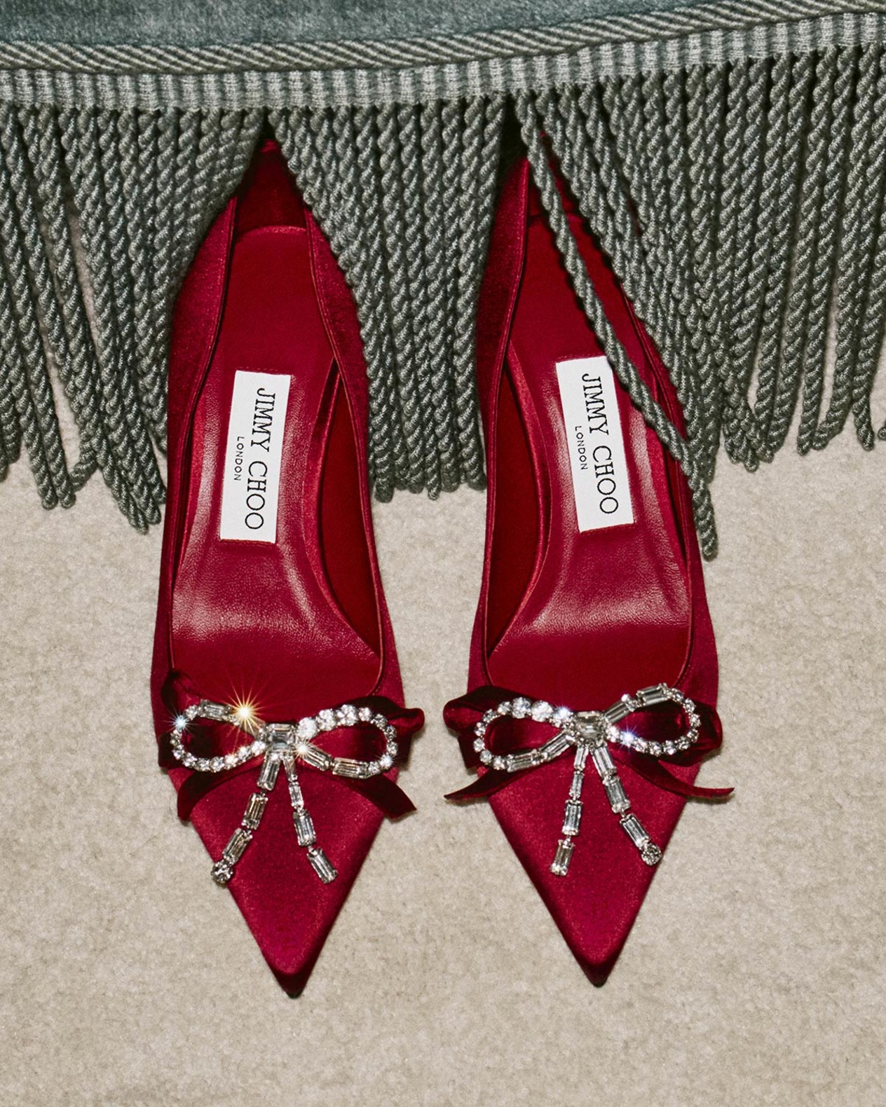 A pair of red satin pumps adorned with crystal bow embellishments, styled beneath a fringed throw.