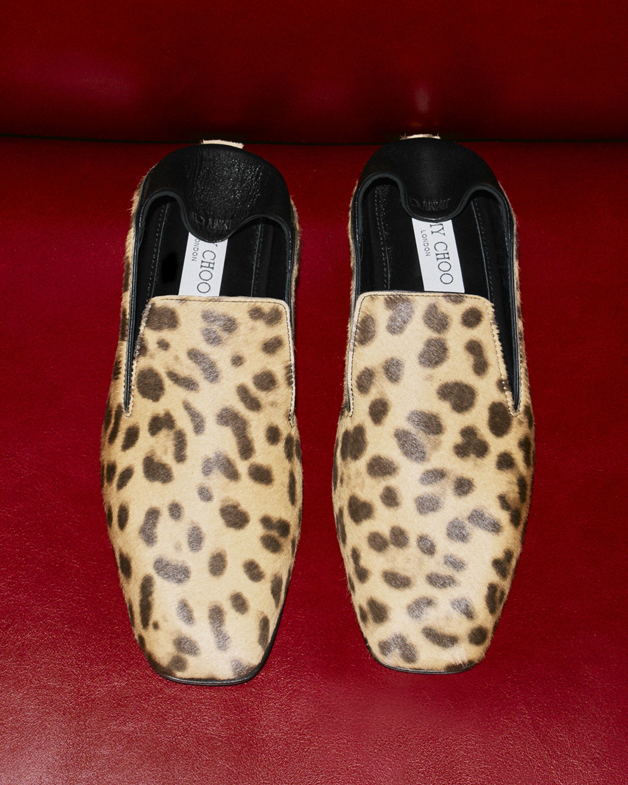 Leopard-print slip-on slippers with square toes and black leather trim, displayed against a bold red leather backdrop.