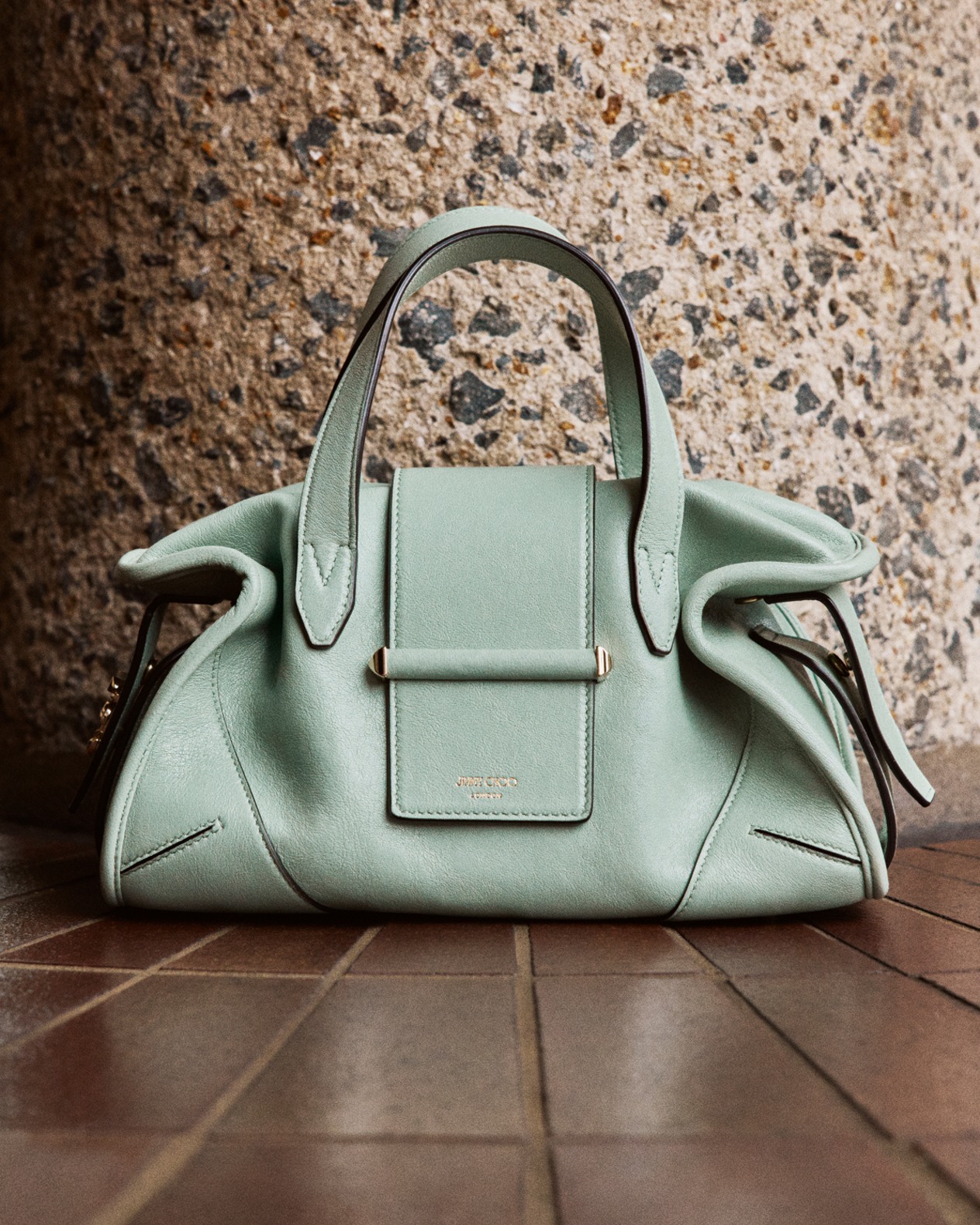 Mint green top-handle handbag with structured silhouette placed on a brick floor against a textured stone wall.	