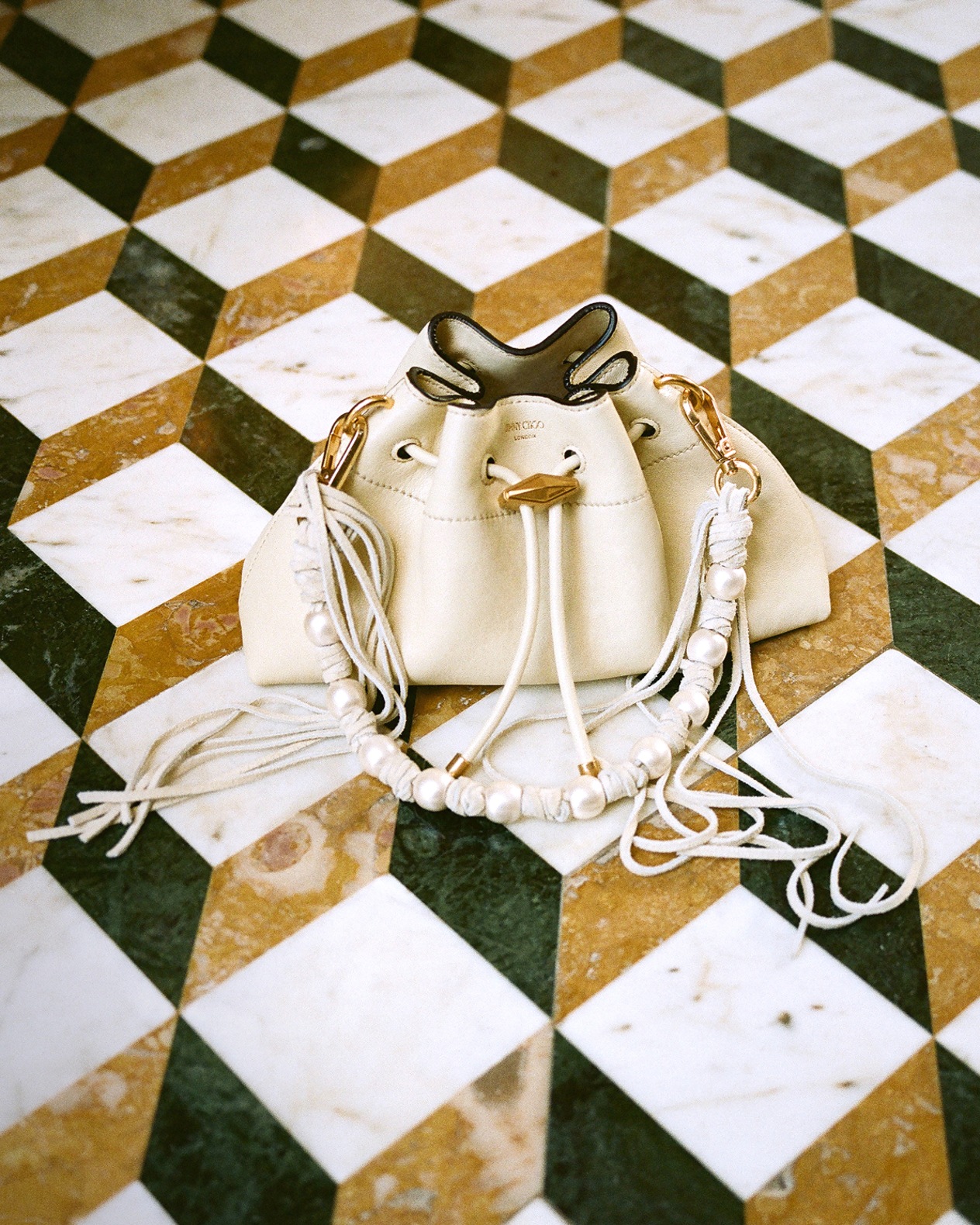 Beige drawstring shoulder bag with dark brown floral embroidery, gold hardware and circular top handle, displayed on a gold door handle against a paneled wall.