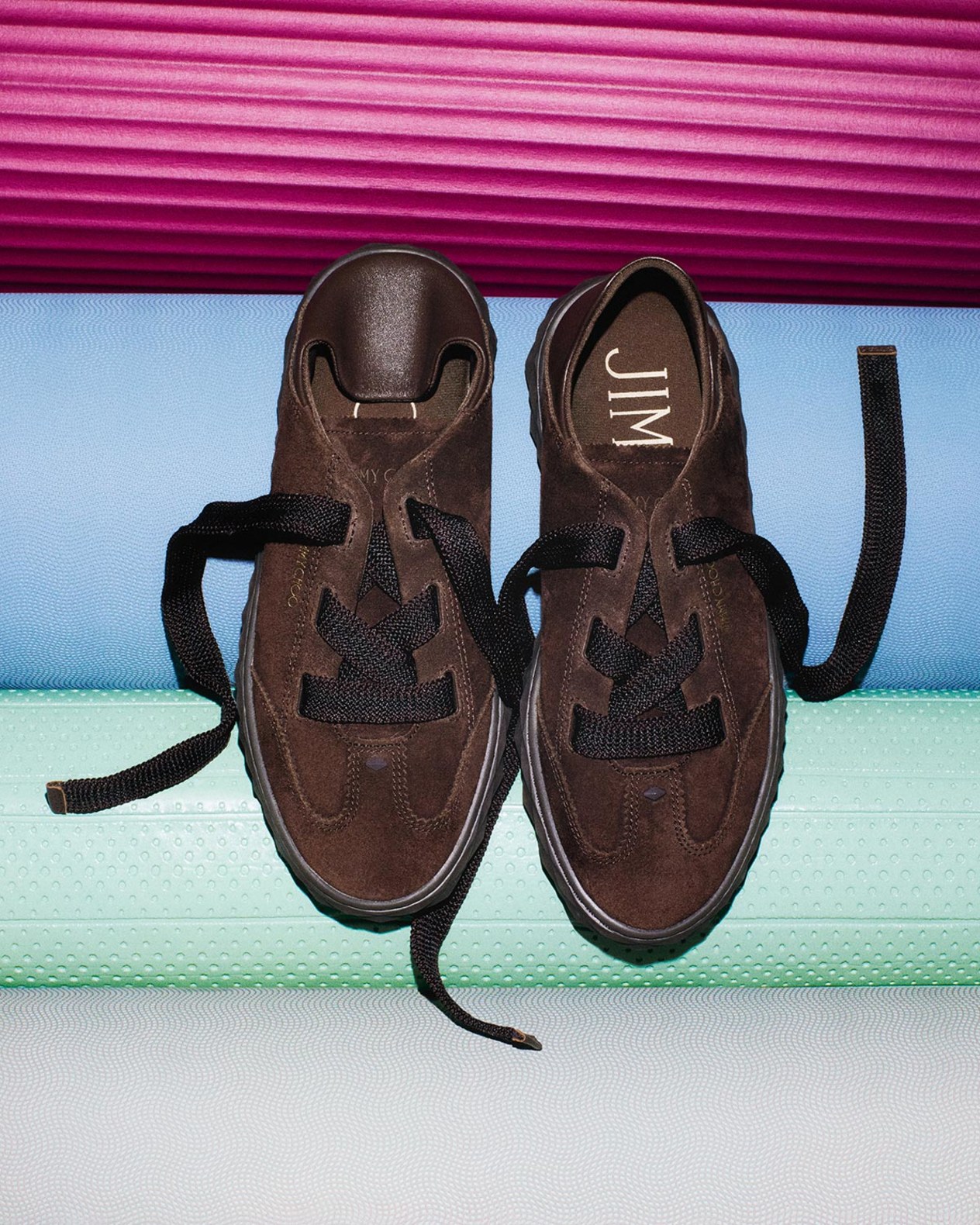 Pair of brown suede trainers with black laces, displayed against a gradient pink, blue and green backdrop.