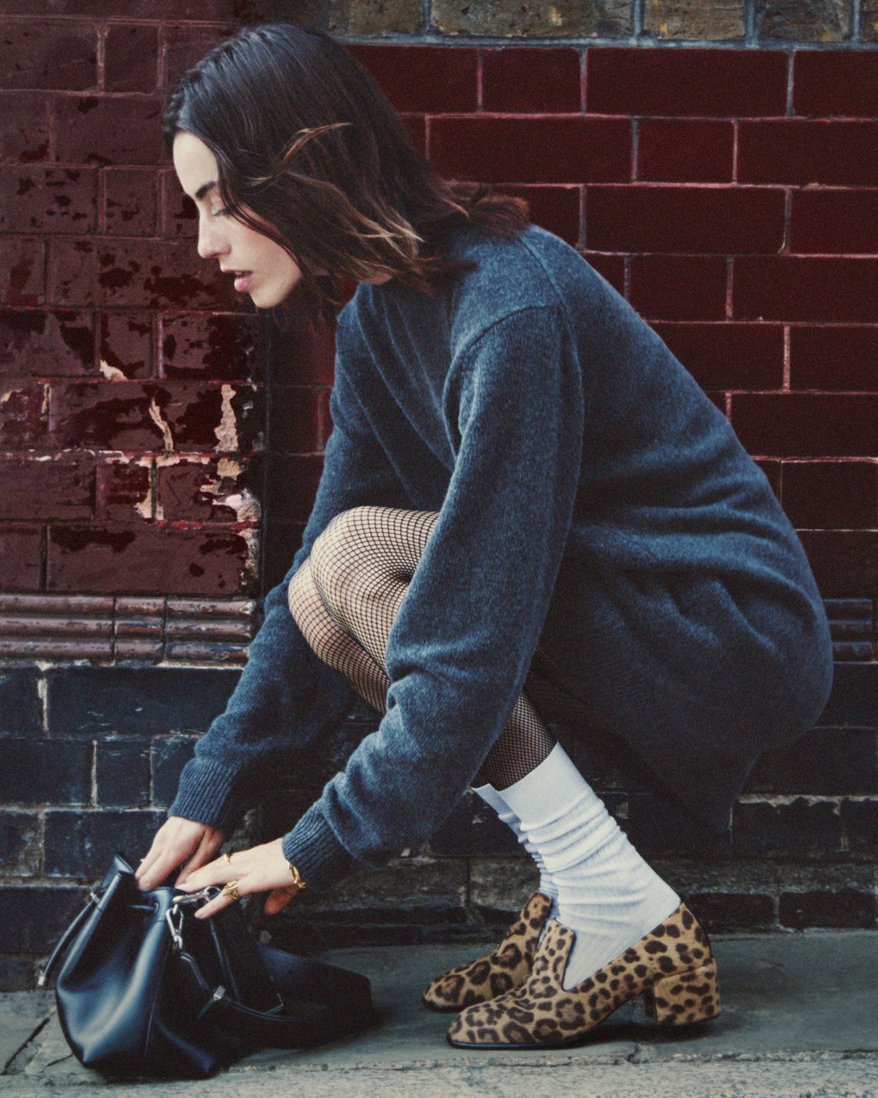 Woman, wearing leopard print heeled Eliot slippers styled with white socks, crouching to look through her bag, styled in a dark oversized sweater and photographed against a brick wall.
