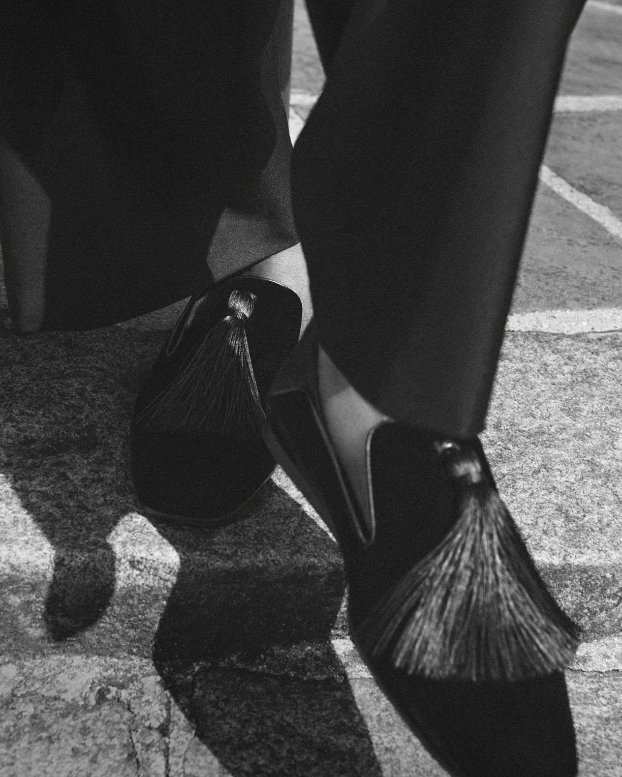Close-up of men’s black velvet Eliot slippers with large tassels, styled with tailored trousers, photographed on a city pavement.