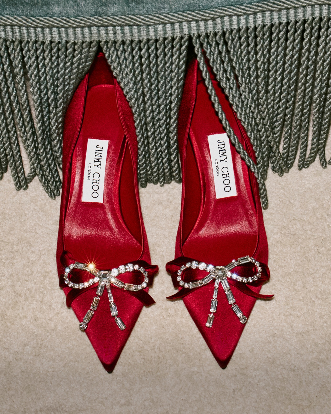 A pair of red satin pumps adorned with crystal bow embellishments, styled beneath a fringed throw.