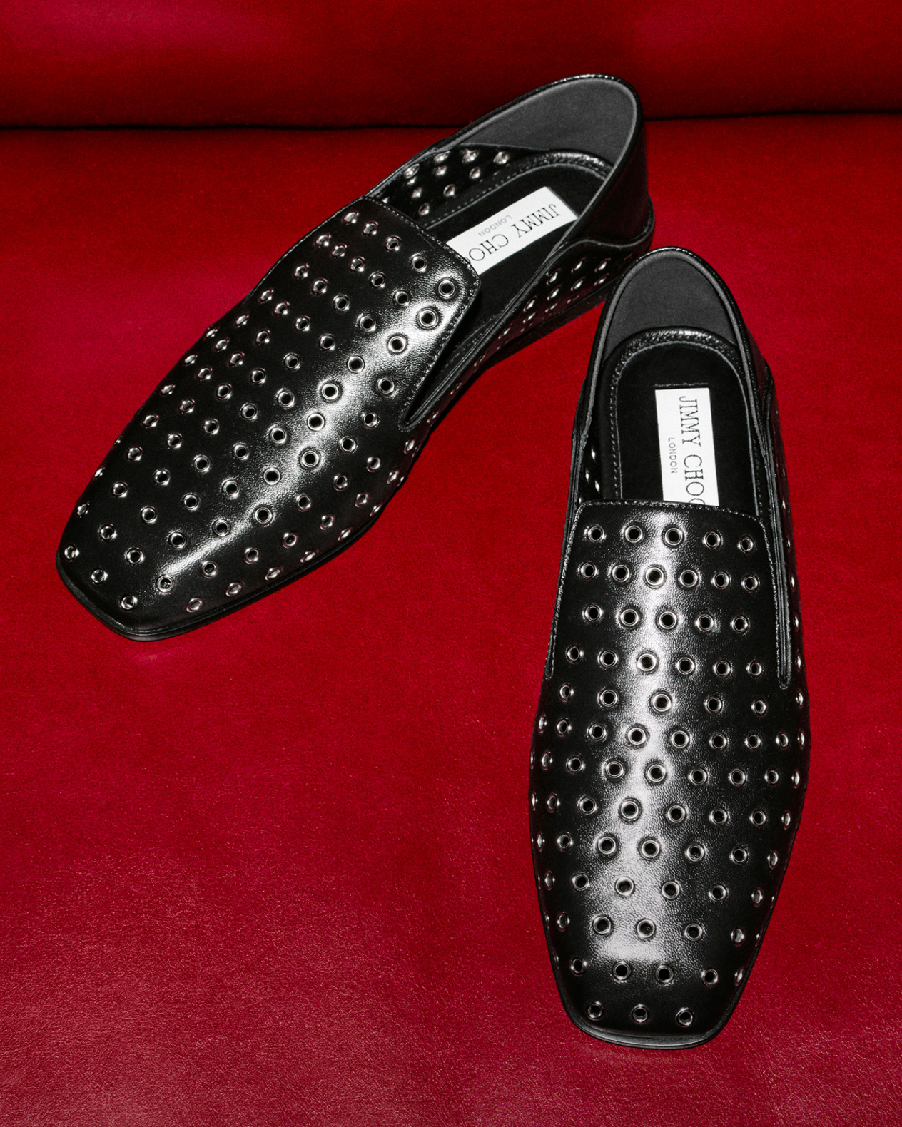 Black eyelet Eliot slipper shoes, displayed against a bold red leather backdrop.