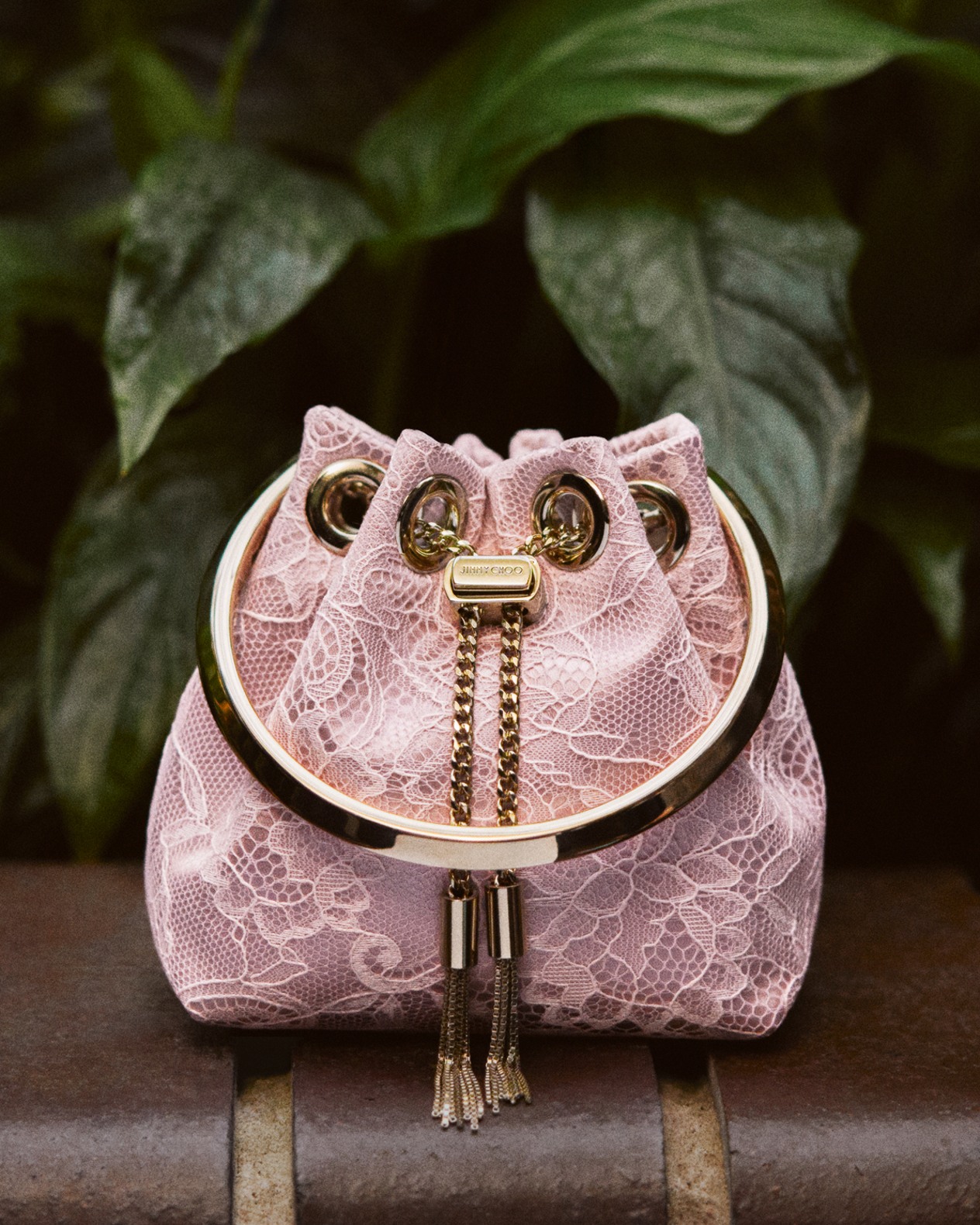 Pink lace drawstring mini bag with gold hardware and tassel details, placed on a stone ledge against a leafy green background.