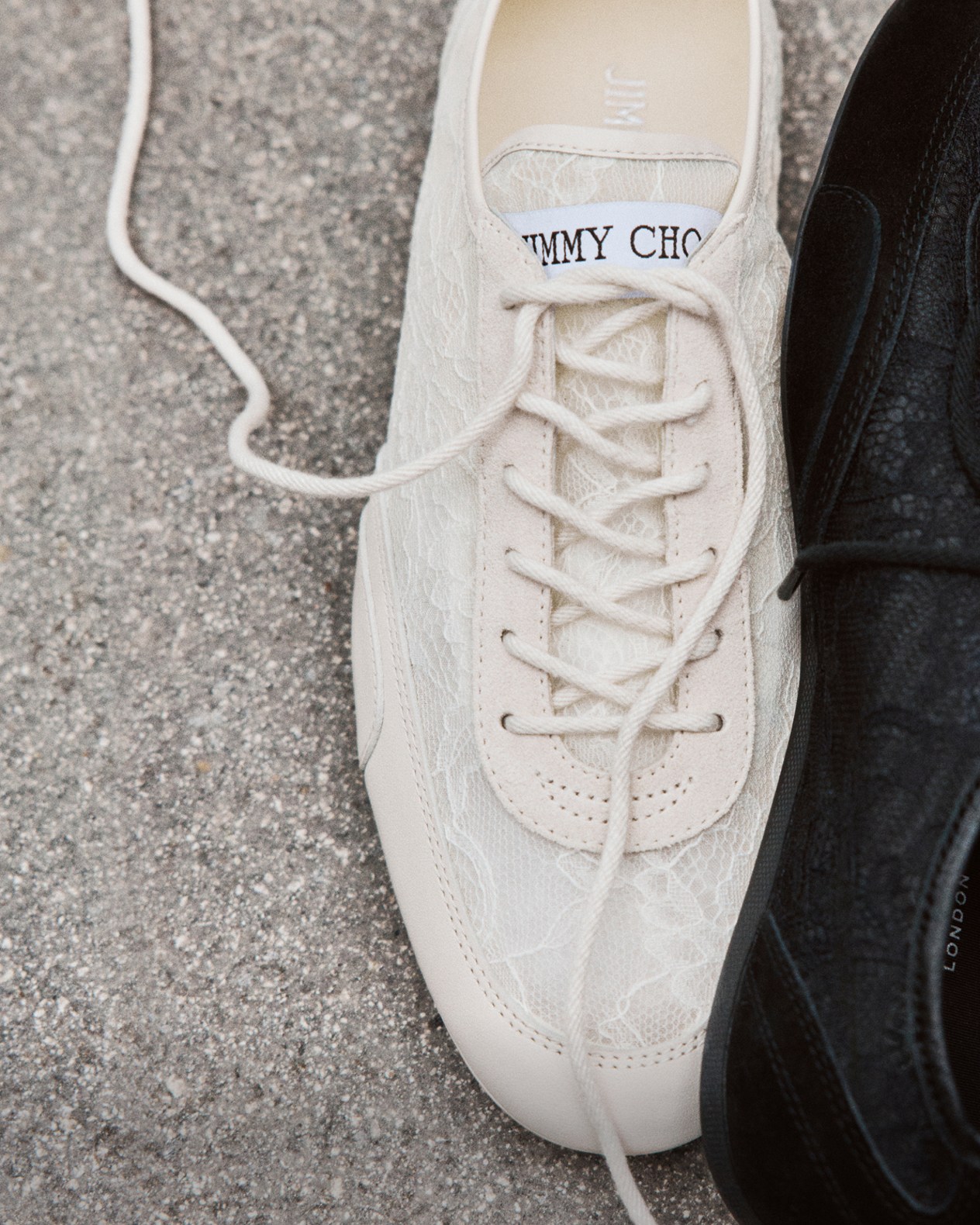 One cream and one black low-top lace-up trainer placed side by side on a grey concrete surface.