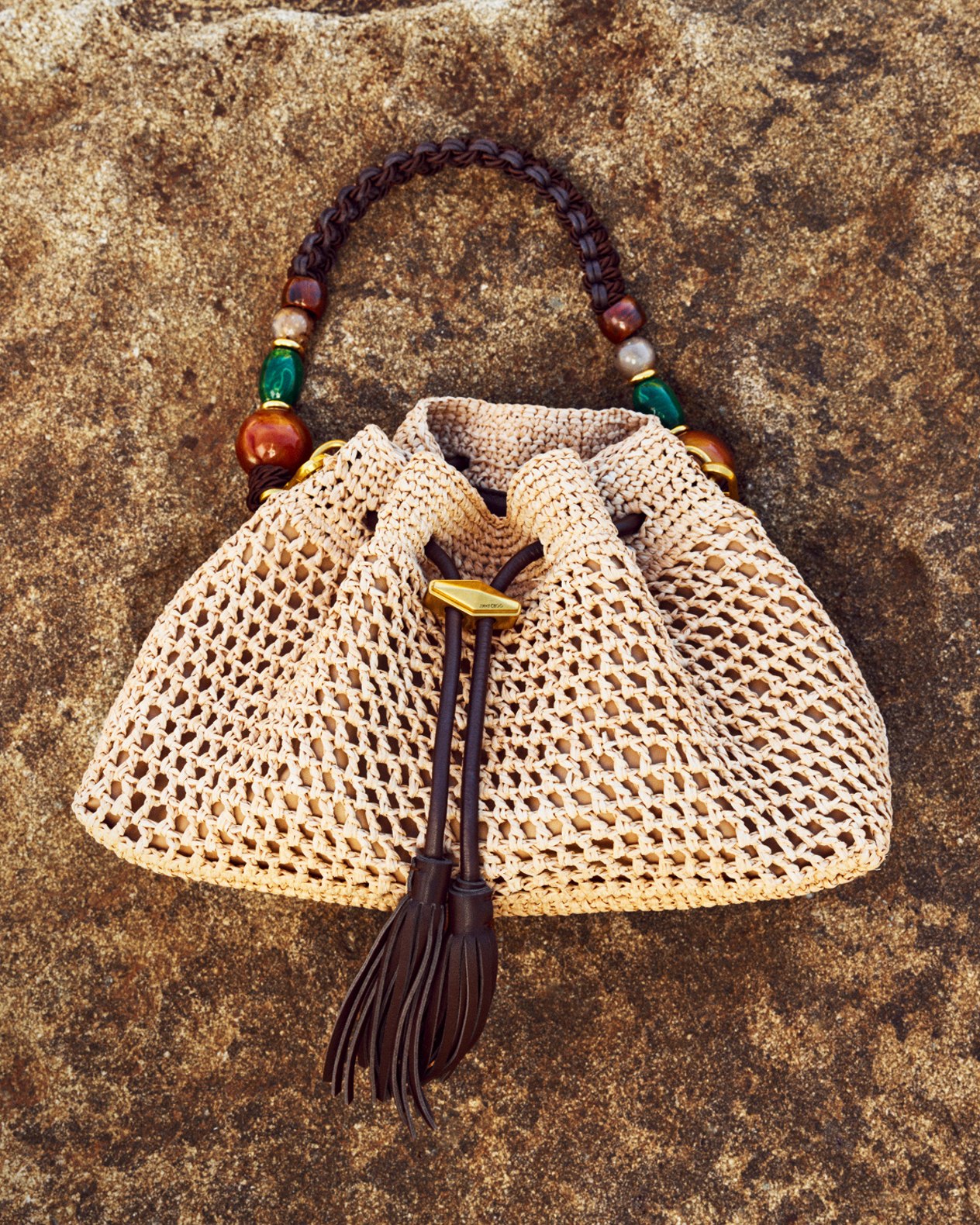 Beige flat sandals with textured fringe straps placed on rocky ground.