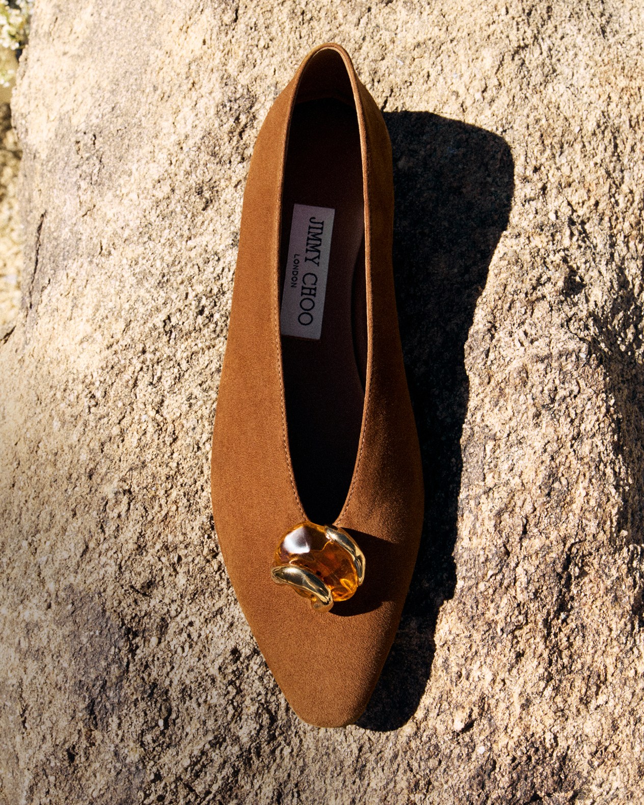 Brown suede flat shoe with a sculptural gold stone embellishment displayed on a textured stone surface.