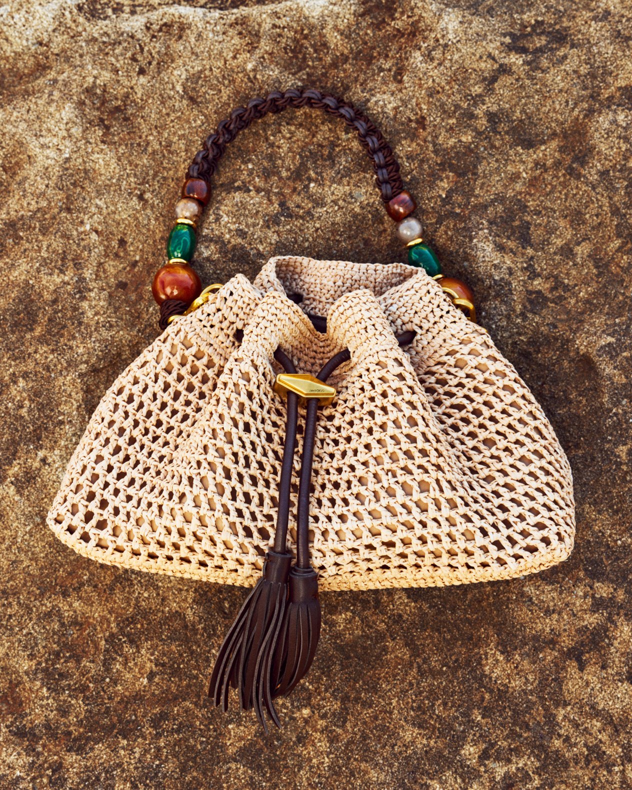 Woven raffia drawstring bag with bead embellishment and tassel detailing displayed against a textured stone background.