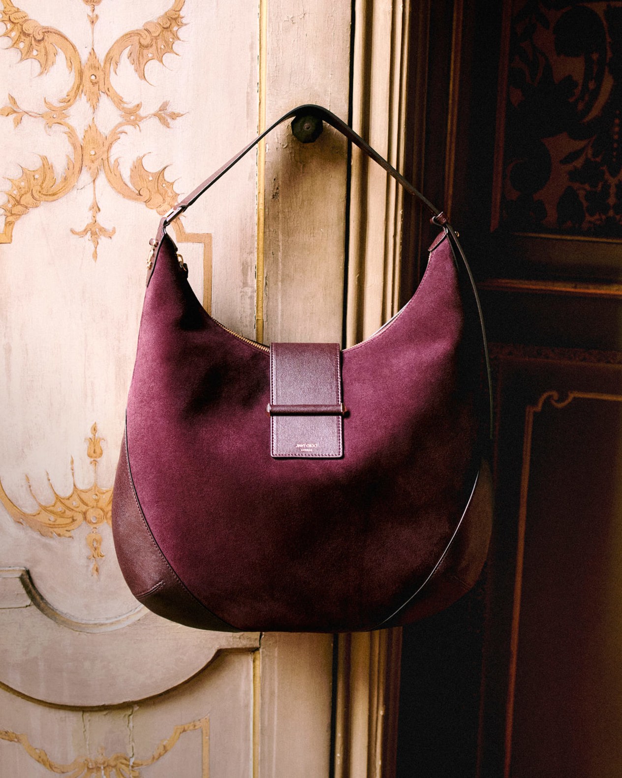 urgundy suede shoulder bag with a leather strap hanging from an ornate doorway.