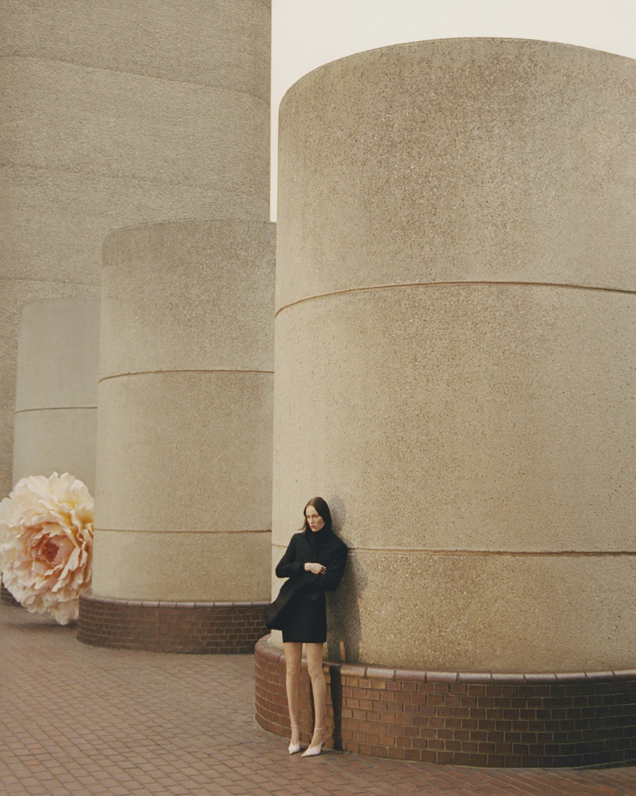 Model standing beside large concrete columns with an oversized flower installation visible in the background.