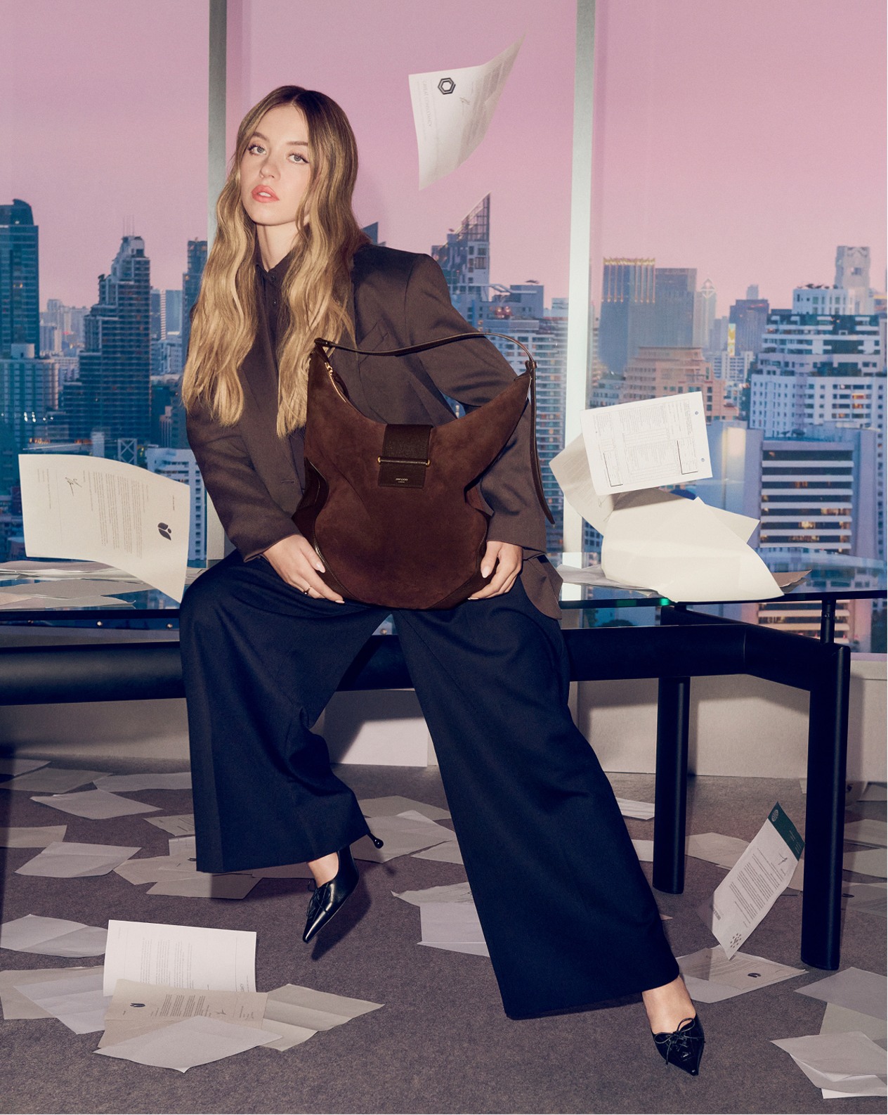 Sydney Sweeney wearing wide-leg trousers and the Scarlett pump, holding the Bar Hobo Large bag in a city office setting with scattered papers and skyscraper backdrop.