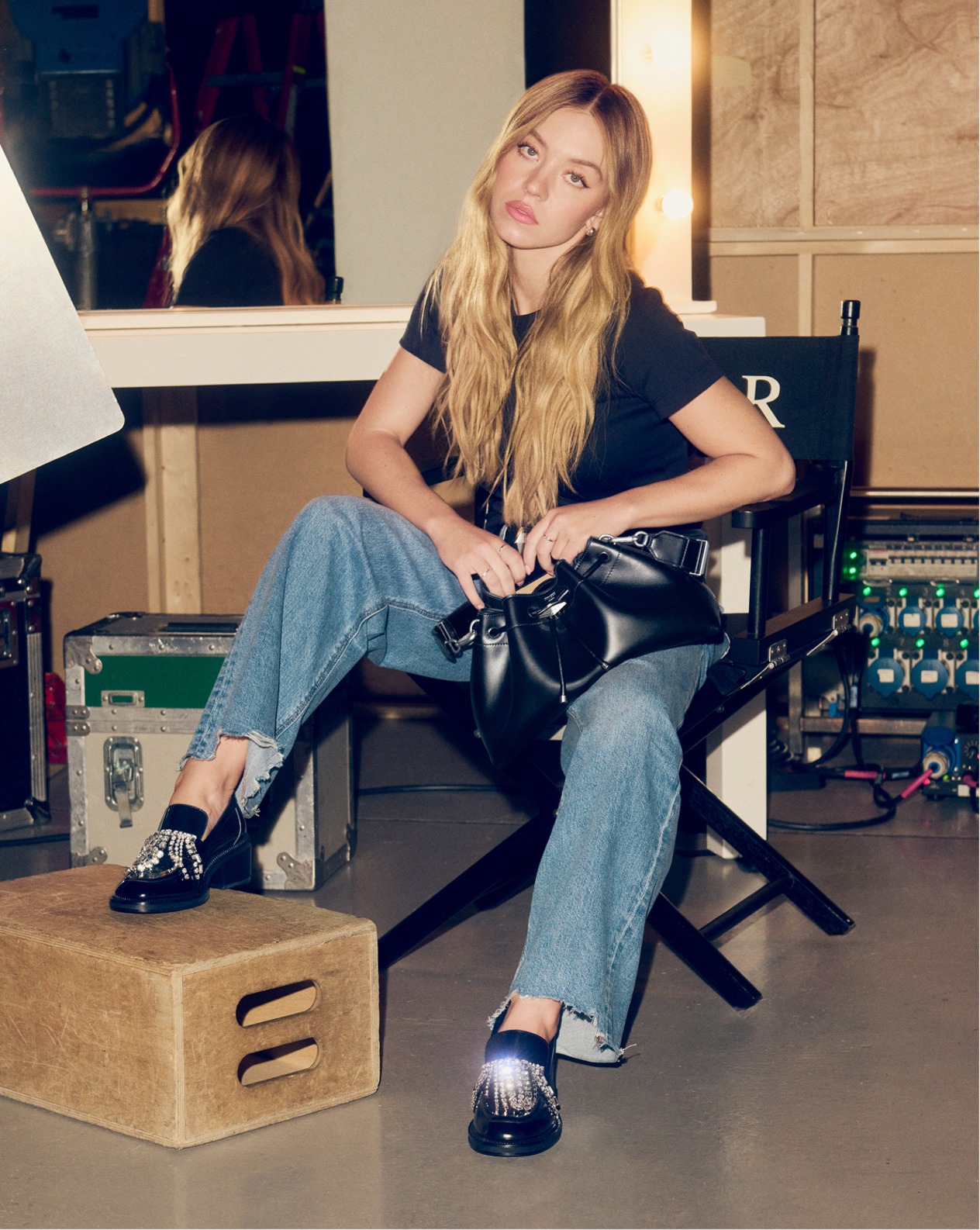 Sydney Sweenet seated backstage in relaxed denim and a black T-shirt, wearing the Tylor loafer with crystal embellishments and holding a black leather Cinch Bag. 