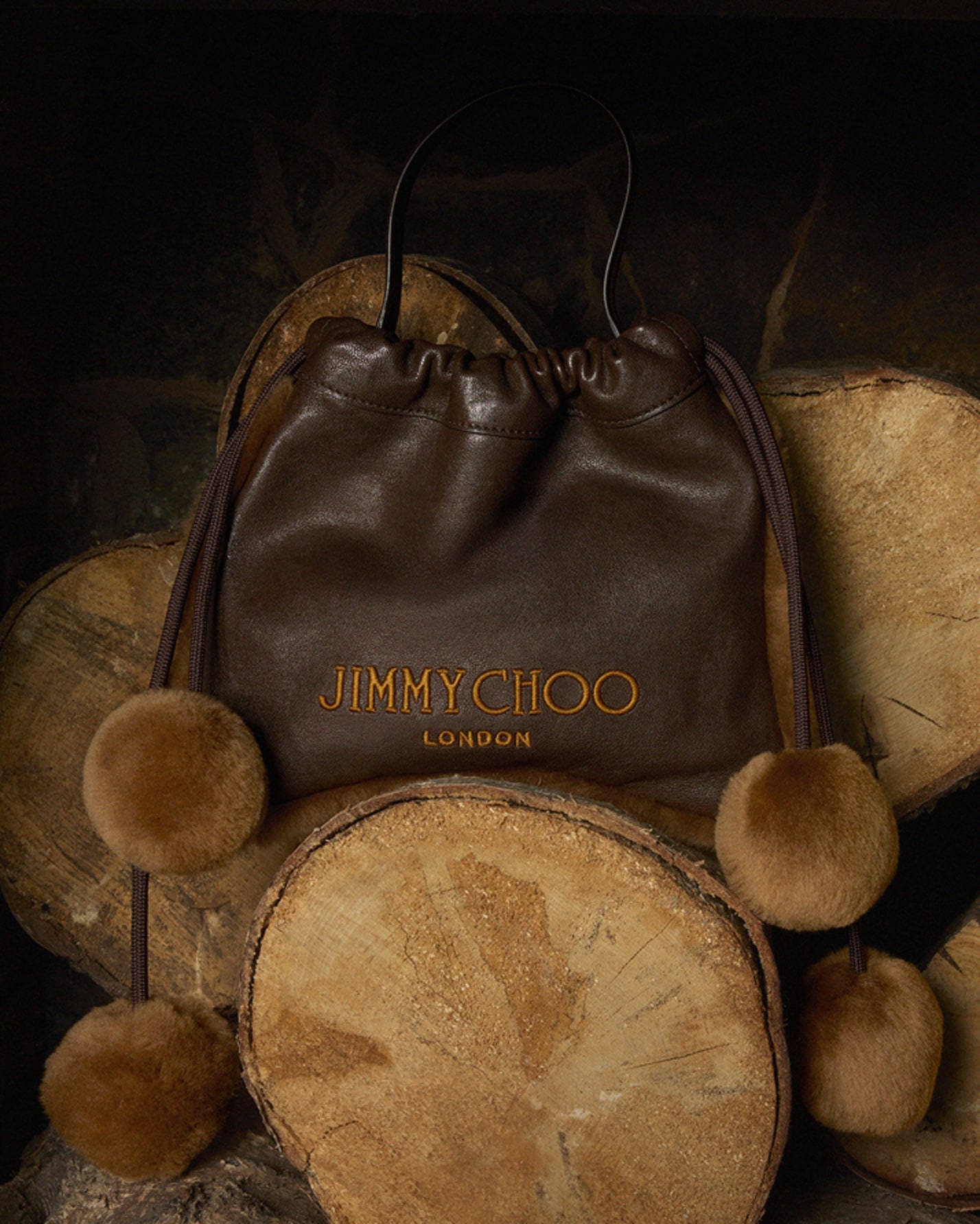 Brown leather drawstring bag with shearling pom-poms and Jimmy Choo branding, displayed among stacked logs.