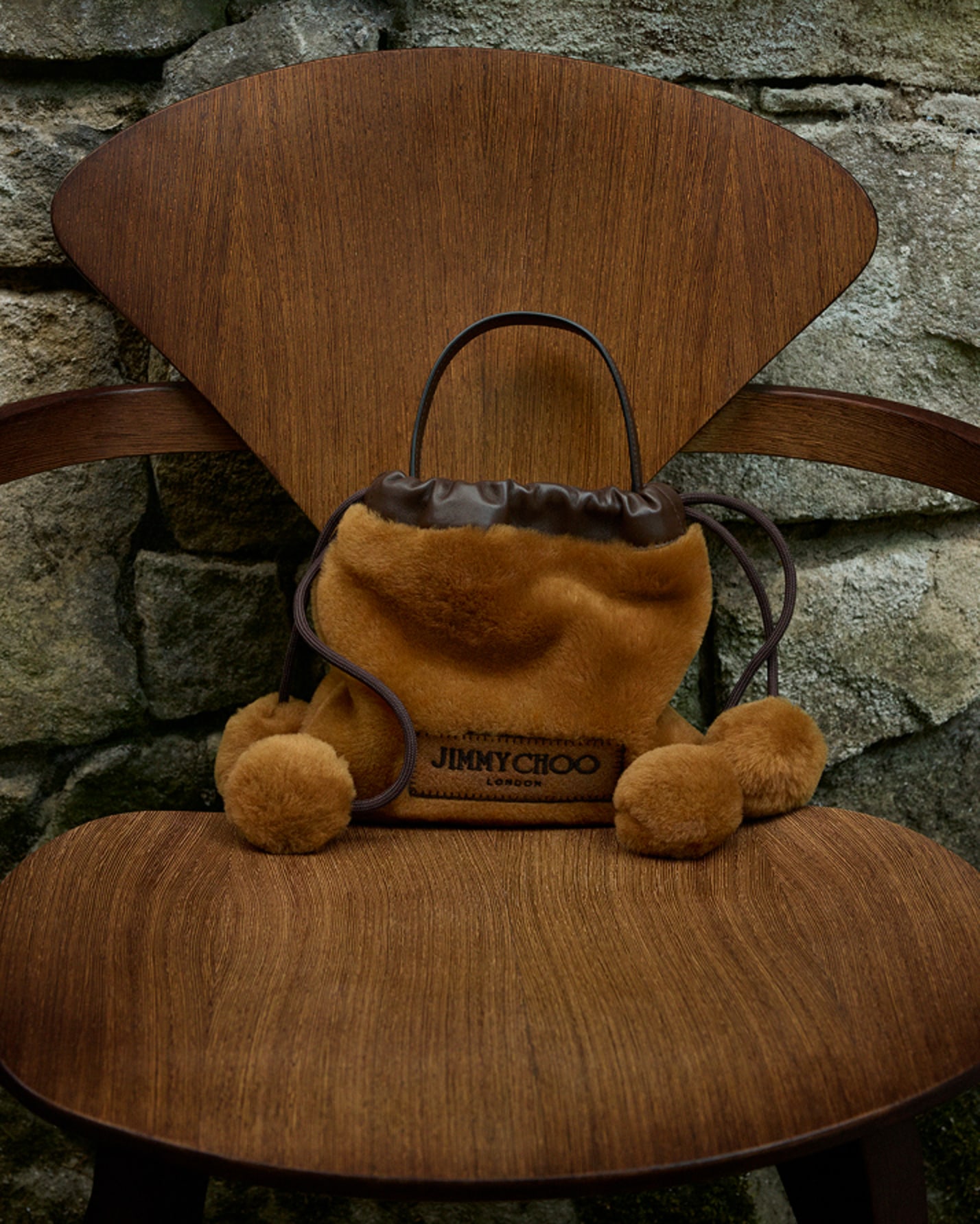 Brown shearling drawstring bag with pom-poms and leather trim, displayed on a wooden chair against a stone wall.