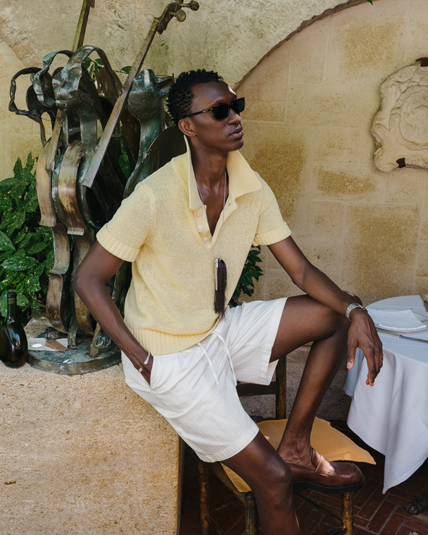 Man seated outdoors wearing sunglasses, a yellow shirt and white trousers.