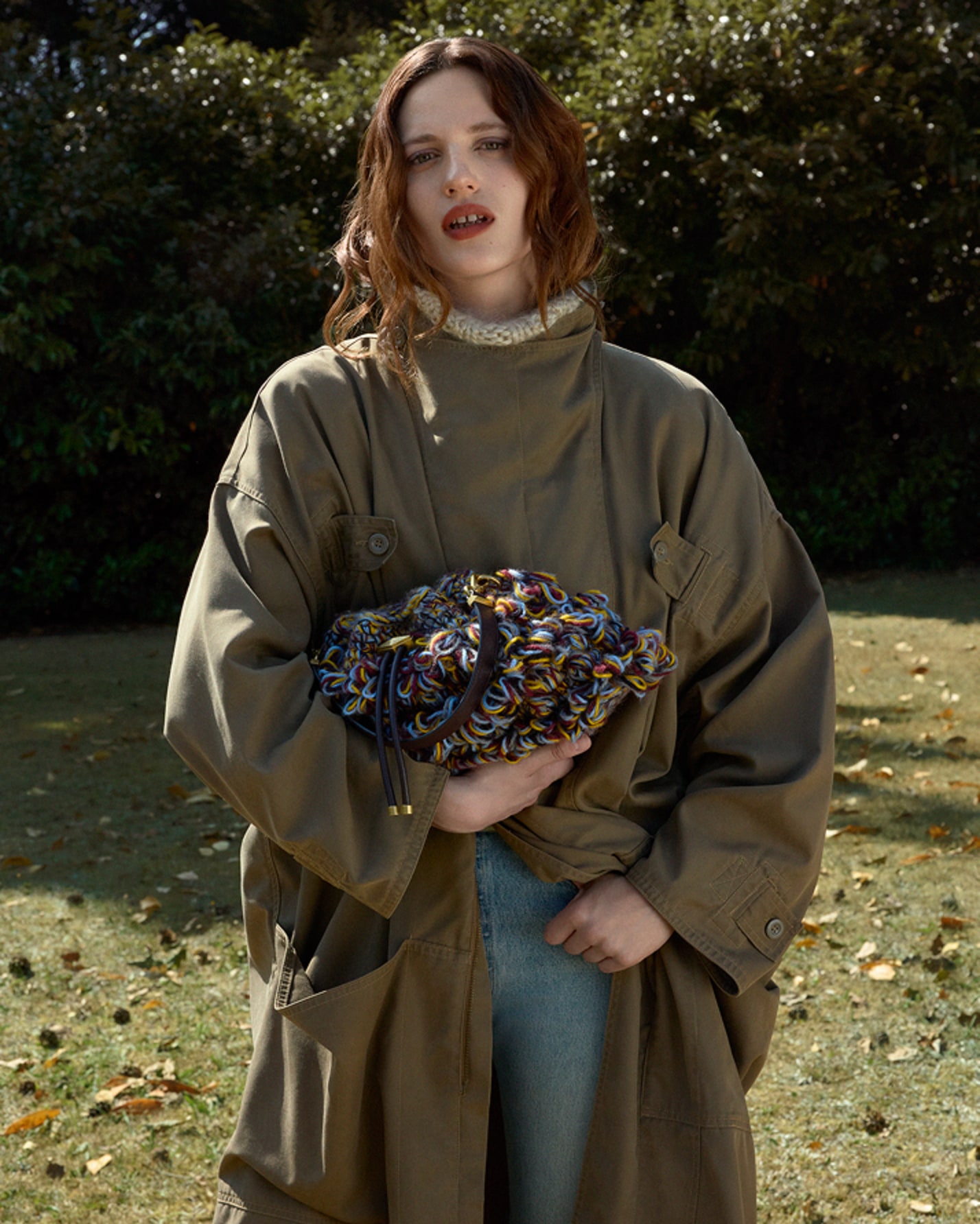Model standing outdoors in a long khaki coat and light-wash jeans, holding a multicoloured woven knit bag.