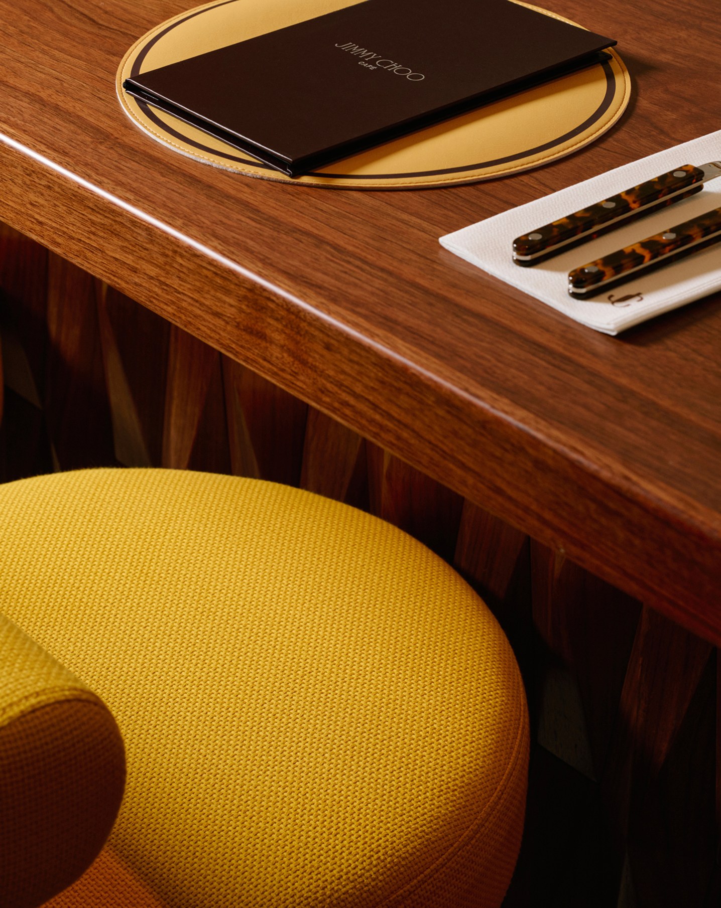 Close-up of a caf&eacute; table with yellow seating and a wooden tabletop holding a black menu and cutlery.