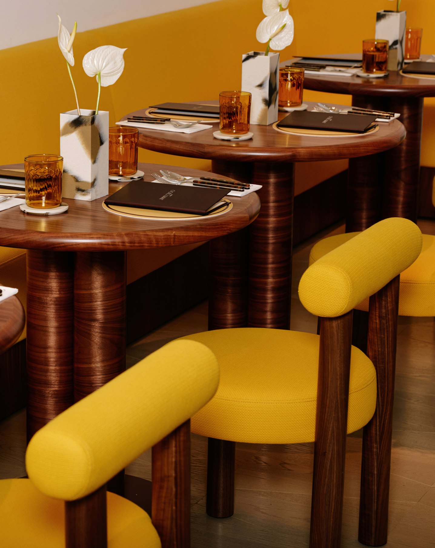  黄色と木目調のインテリアの中で、メニュー、テーブルウェア、花がセットされたカフェテーブルと椅子のクローズアップ。