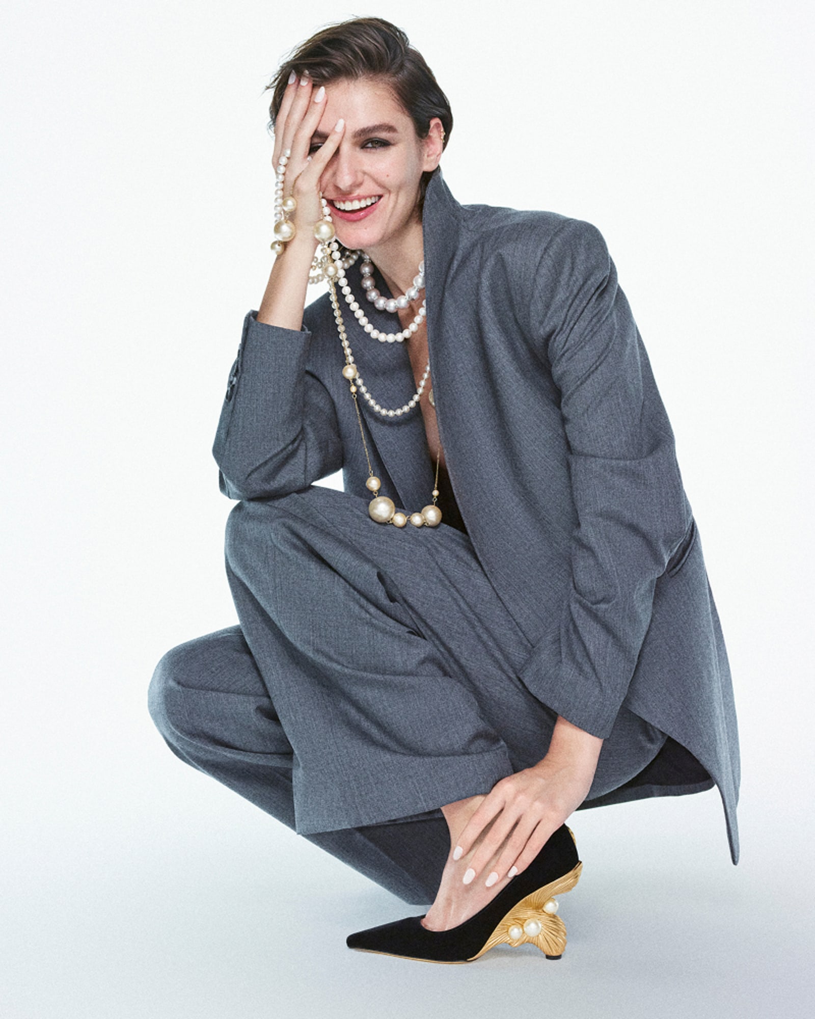 Model crouching in a grey oversized suit and layered pearl necklaces, wearing black pointed-toe pumps with sculptural gold heels and pearl detailing.