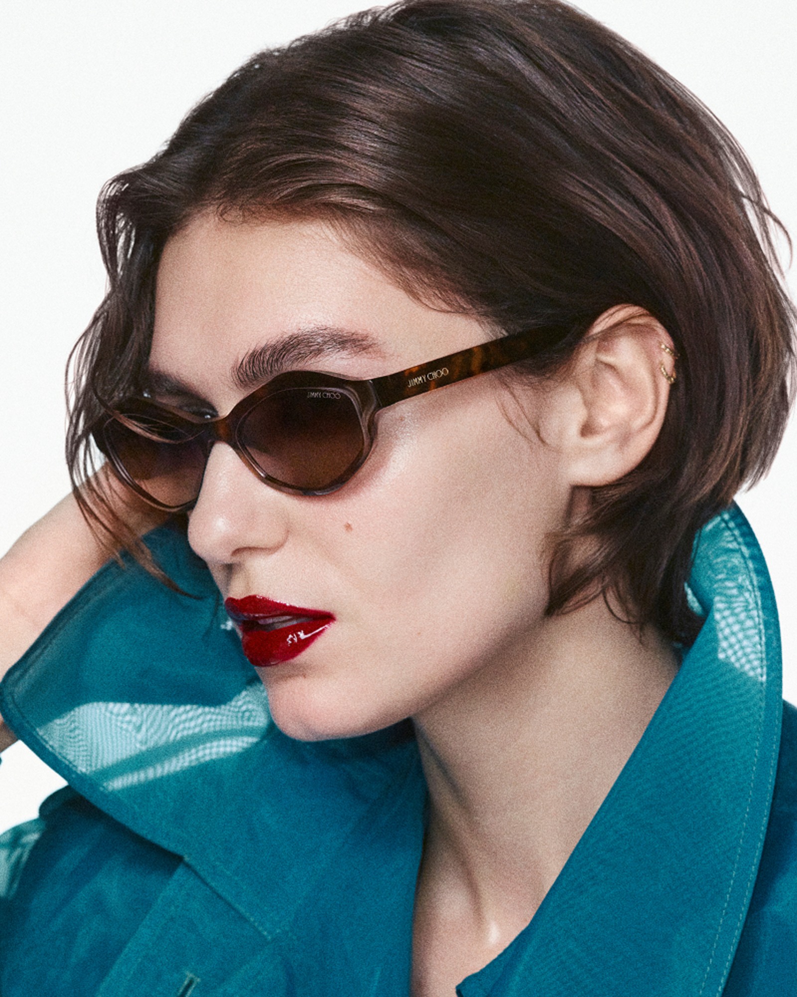 Close-up of a model with short brown hair wearing oversized tortoiseshell sunglasses, red lipstick and a teal blouse with a sheer collar against a white background.